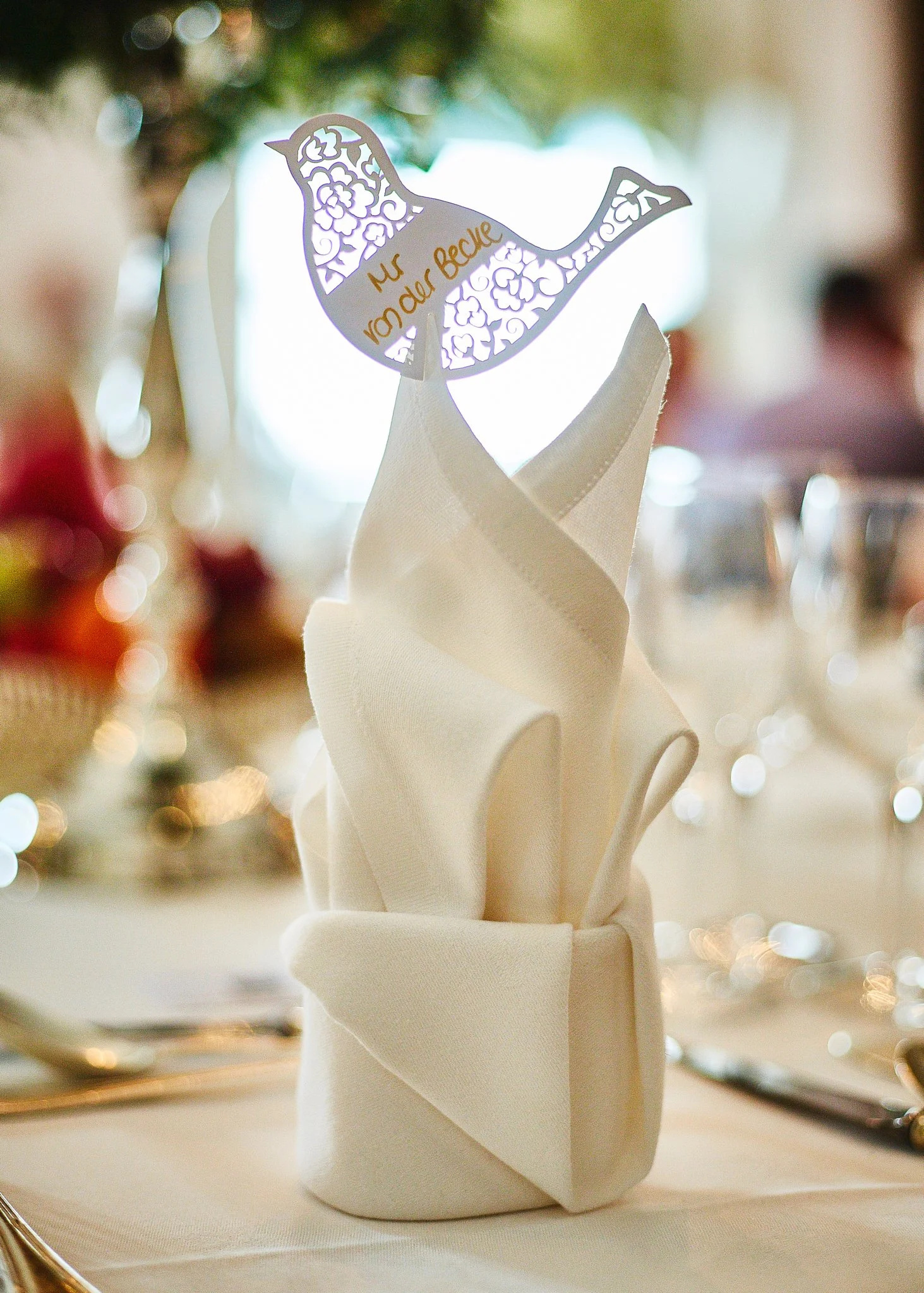A white napkin folded into a tall, elegant shape with a decorative bird-shaped paper cutout on top, featuring floral patterns and the words "inonderve" in gold.