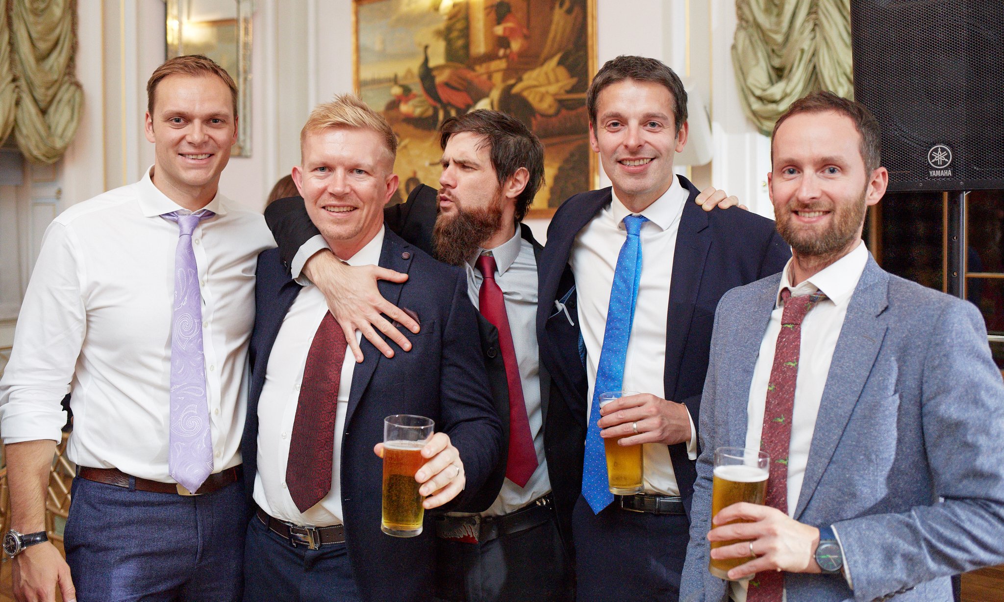 A group of six men in formal attire, smiling and holding drinks at a social event or party in an elegant room with artwork and curtains in the background.