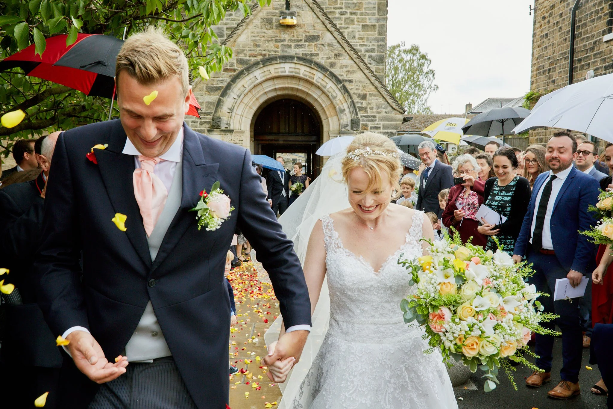 A newlywed couple holding hands and walking down the aisle after their wedding ceremony outside a church, surrounded by family and friends, with flower petals on the ground and umbrellas overhead.
