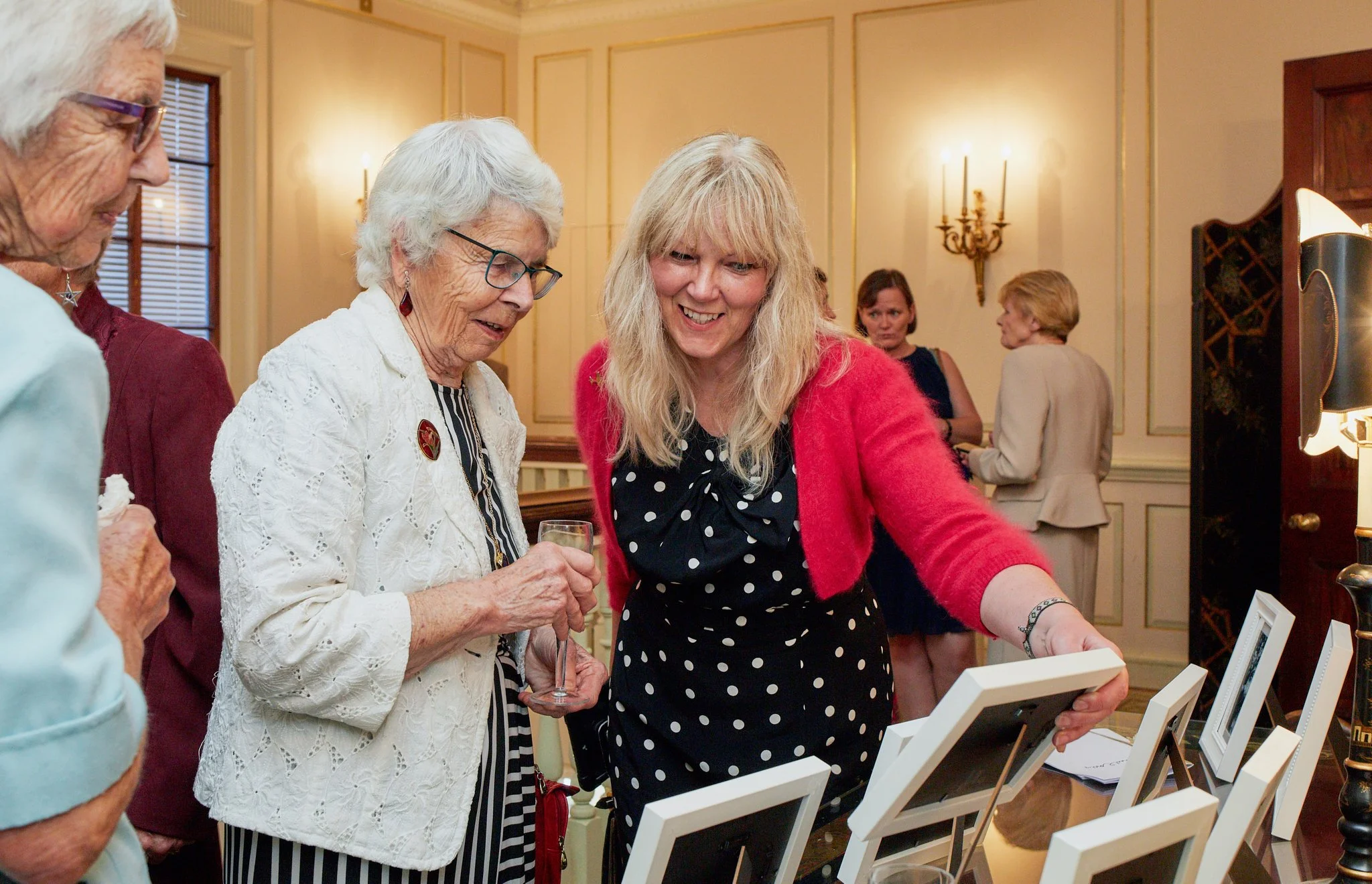 Women and elderly women gathered in a room, looking at framed photographs or artwork on display, with warm lighting and elegant decor.