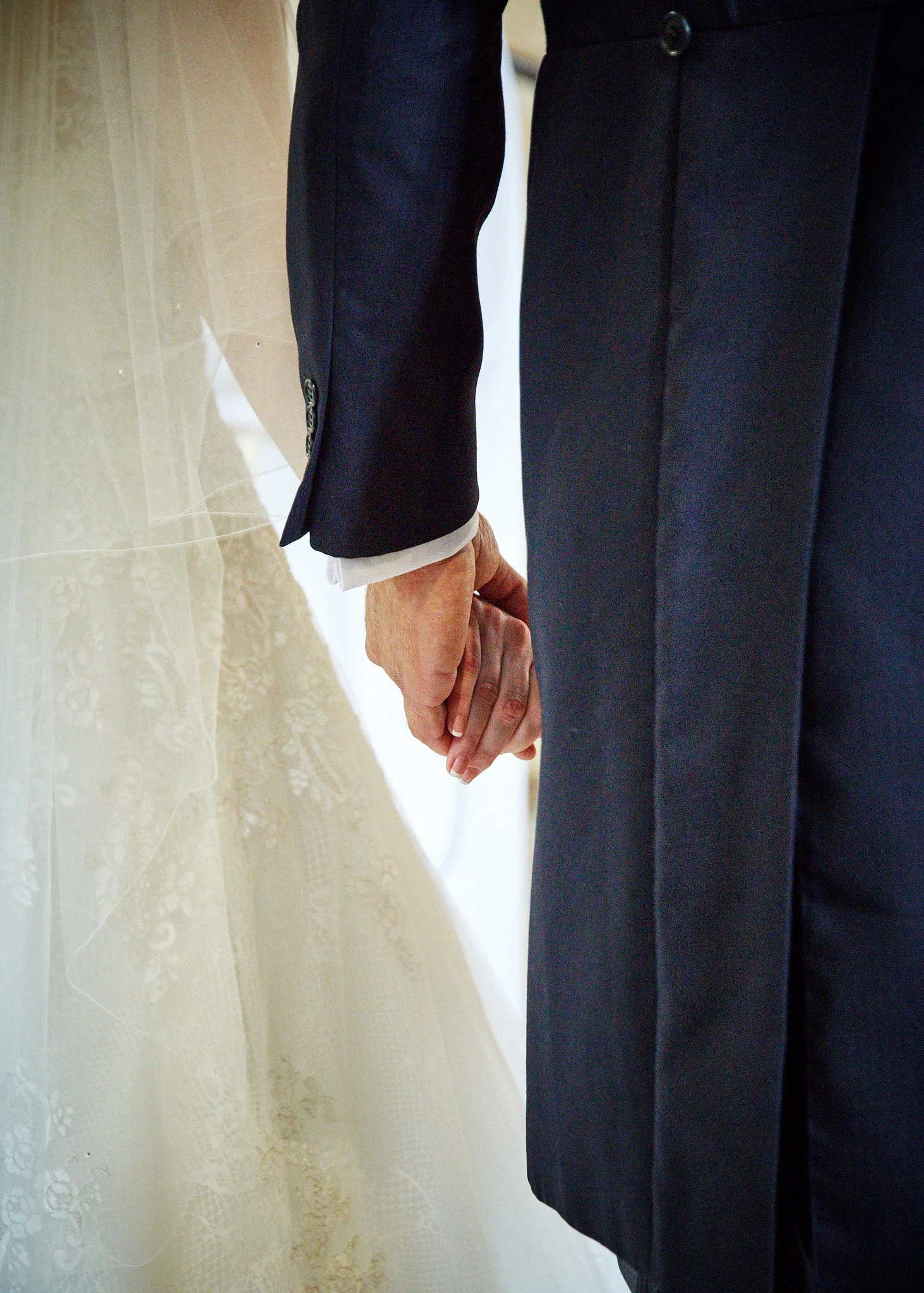 Close-up of two people holding hands, one wearing a wedding dress with lace details and the other in a dark suit with buttoned cuffs.