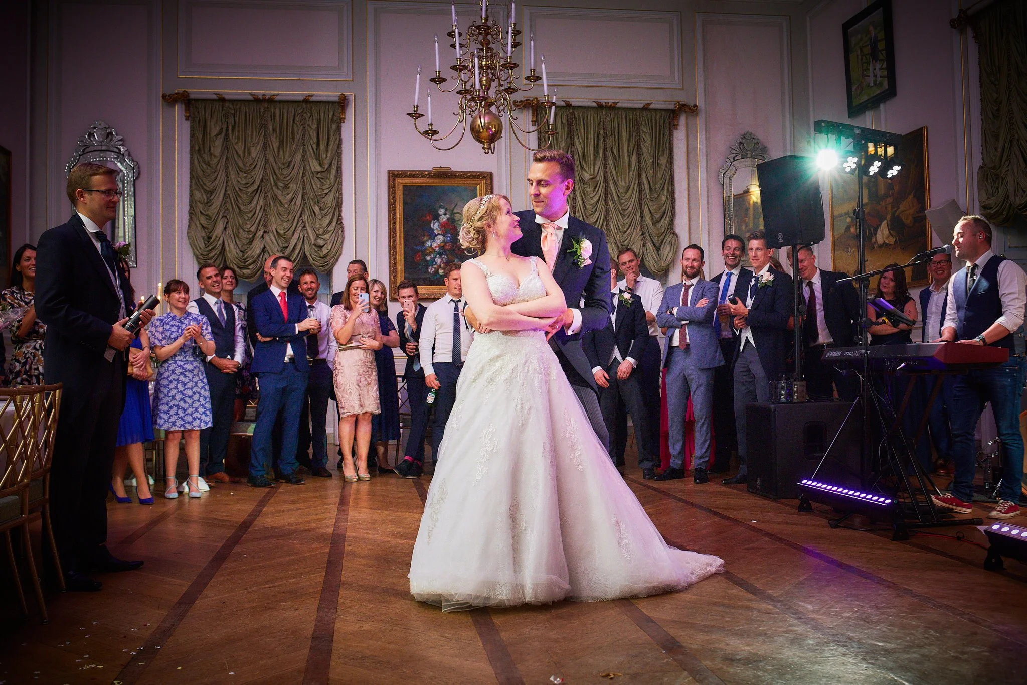 Bride and groom dancing during a wedding reception, surrounded by guests in a grand hall with ornate decorations and gold accents.