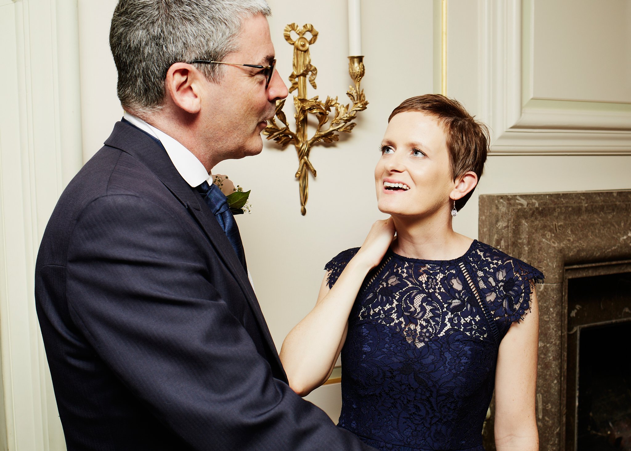 A man and woman are standing close together, looking into each other's eyes. The man is wearing a dark suit, glasses, and has gray hair. The woman is wearing a dark blue lace dress with earrings, and has short brown hair. They are smiling and appear 