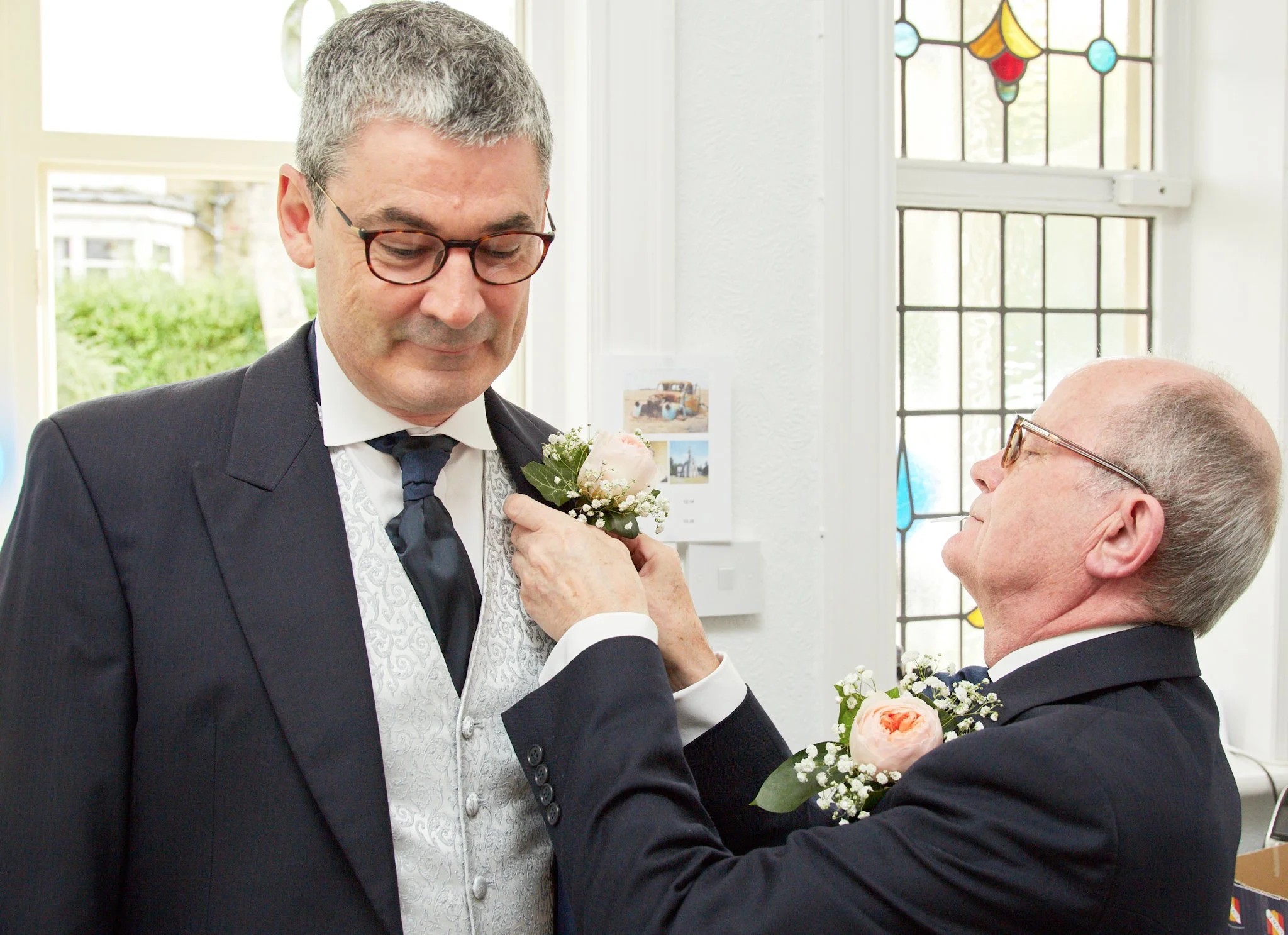 A man in a tuxedo receiving a boutonniere or flower from a woman during a wedding ceremony inside a church with stained glass windows.