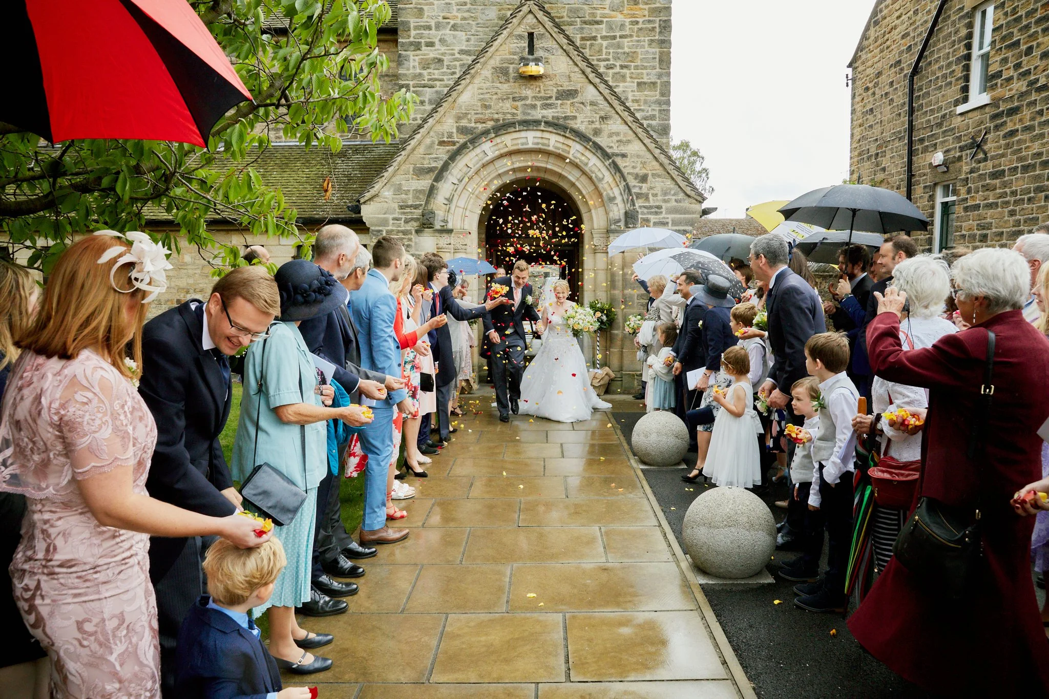 A wedding ceremony outside on a rainy day with guests holding umbrellas, with a bride in a white dress and a groom in a suit, walking away from the church entrance decorated with flowers, as friends and family look on.