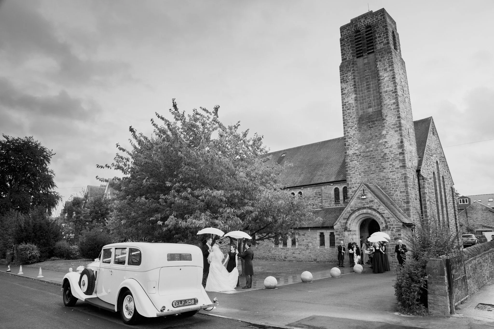 Black and white photo of a church with tall tower, trees in front, and wedding guests outside, including a bride and groom with an escort. Vintage car parked nearby.