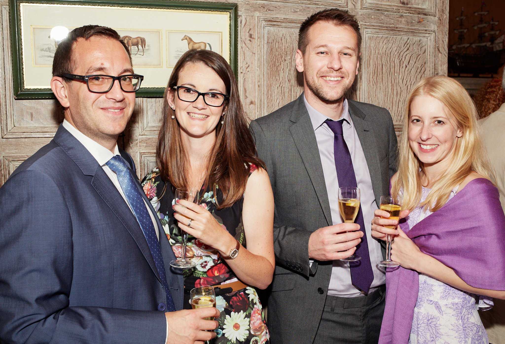 Group of five friends at a celebration, dressed in formal attire, holding glasses of champagne, smiling, against a rustic wooden wall with horse artwork.