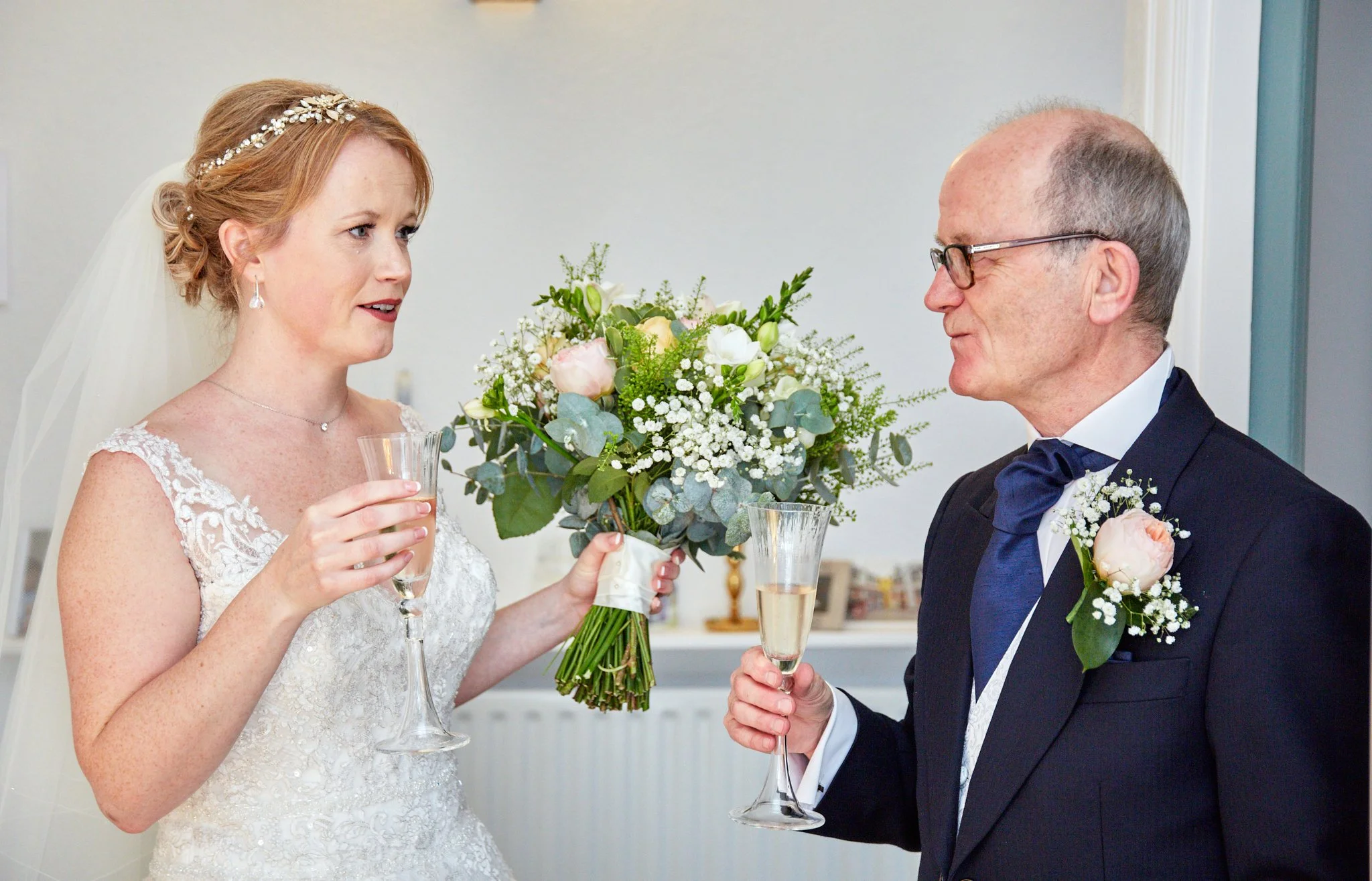 A bride and an older man, possibly her father, sharing a moment at a wedding. The bride has red hair, wearing a wedding dress, and holding a bouquet of flowers. The man is dressed in a tuxedo with a boutonniere. Both are holding champagne glasses and