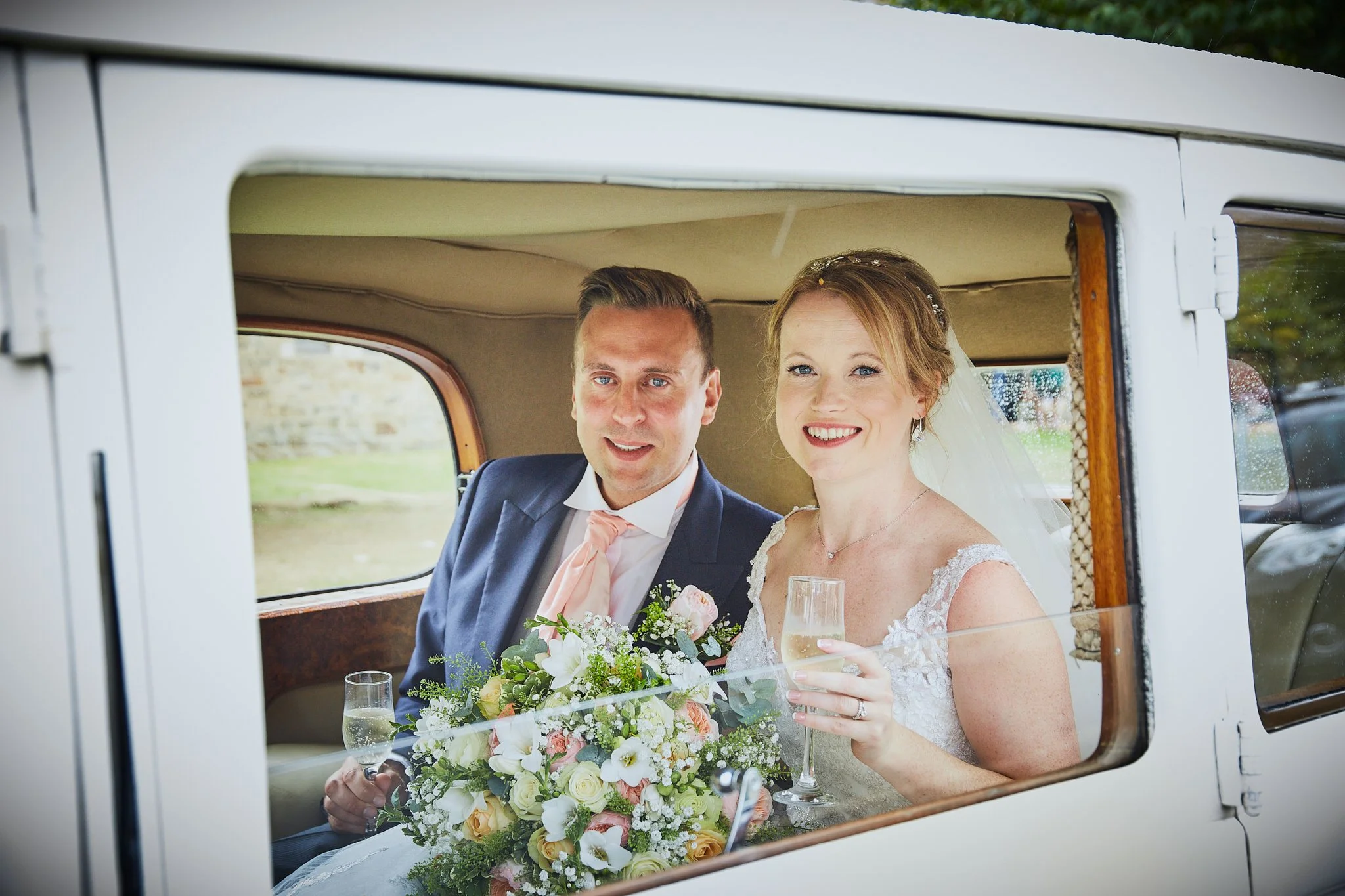 A bride and groom sitting inside a vintage vehicle, smiling and holding glasses, with a bouquet of flowers on the table in front of them.