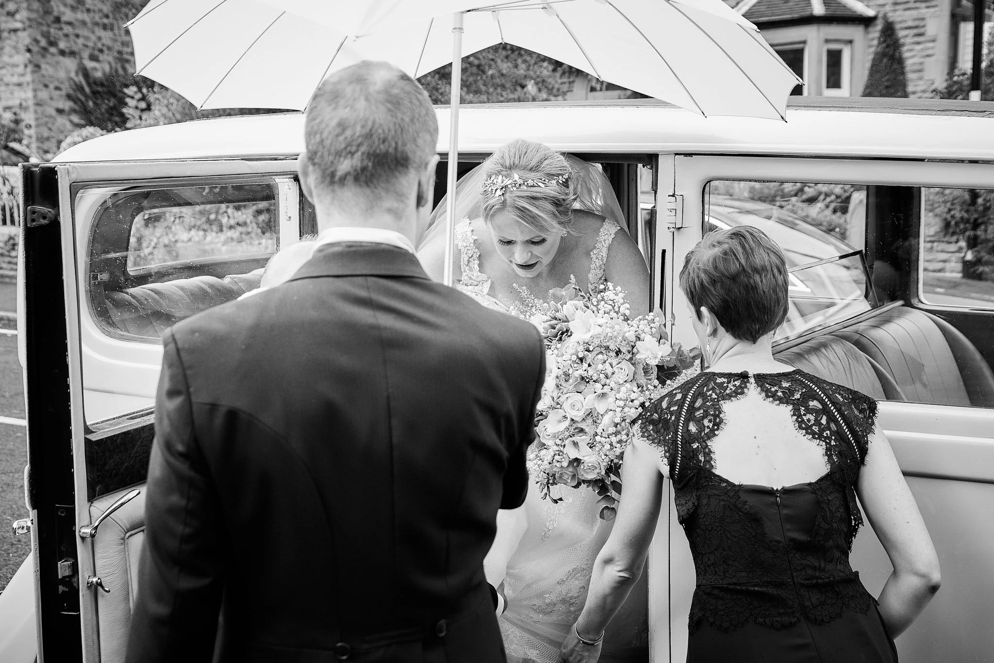A bride exiting a vintage car with the help of a woman, while a man and another woman assist. The bride is holding a bouquet and wearing a lace dress. The scene is set outdoors.