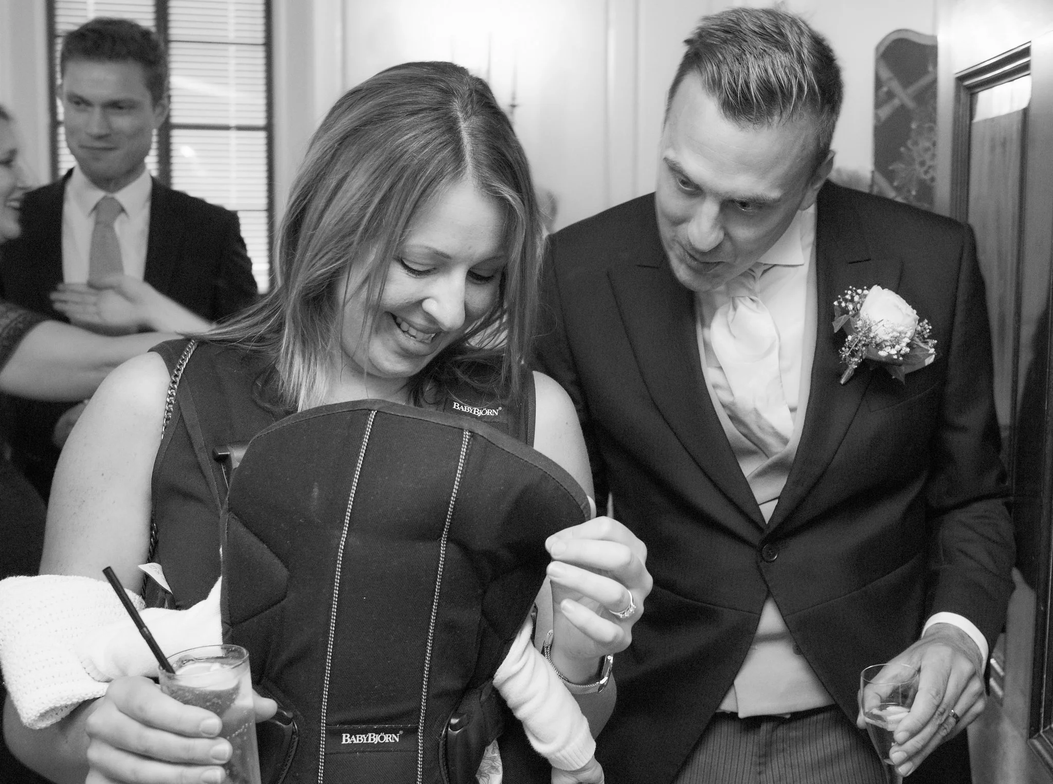 A woman in a dress and a man in a suit are standing together, looking at a phone and smiling. They appear to be at a social event, possibly a wedding, with other people and framed pictures visible in the background.