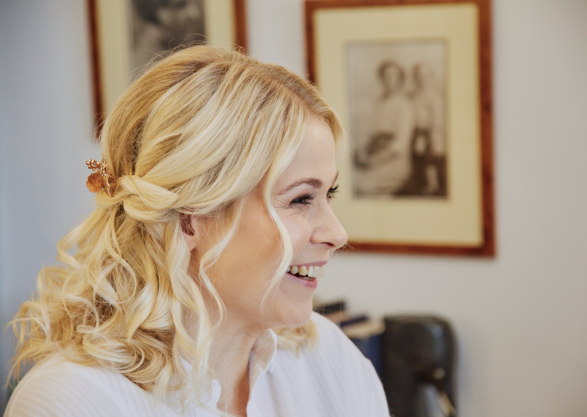 A smiling woman with long, blonde, curly hair styled with a floral hair clip, wearing a white top, in an indoor setting with framed art on the wall behind her.