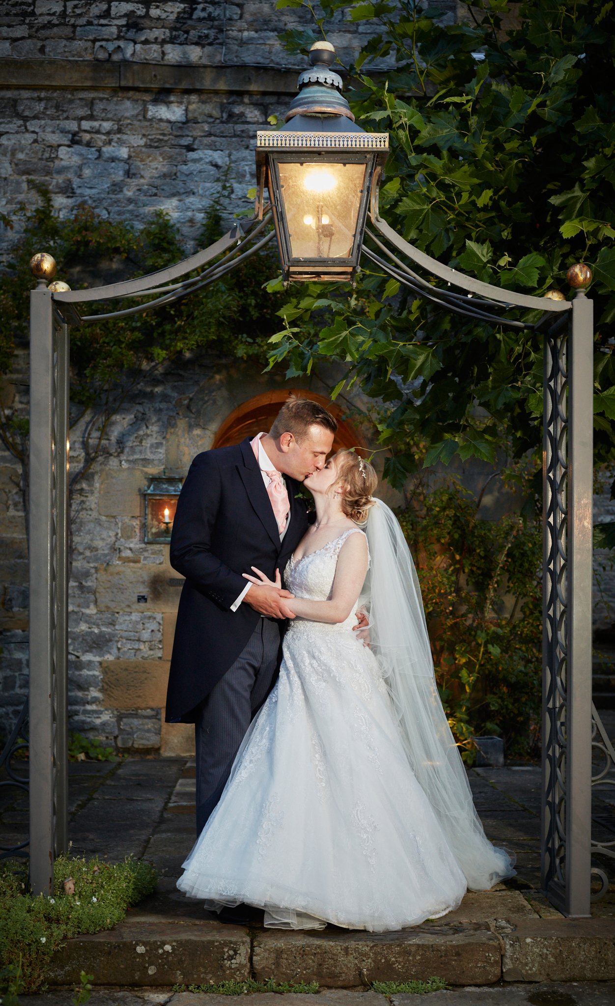 A bride and groom share a kiss under an outdoor lamp archway at dusk, with stone walls and greenery in the background.