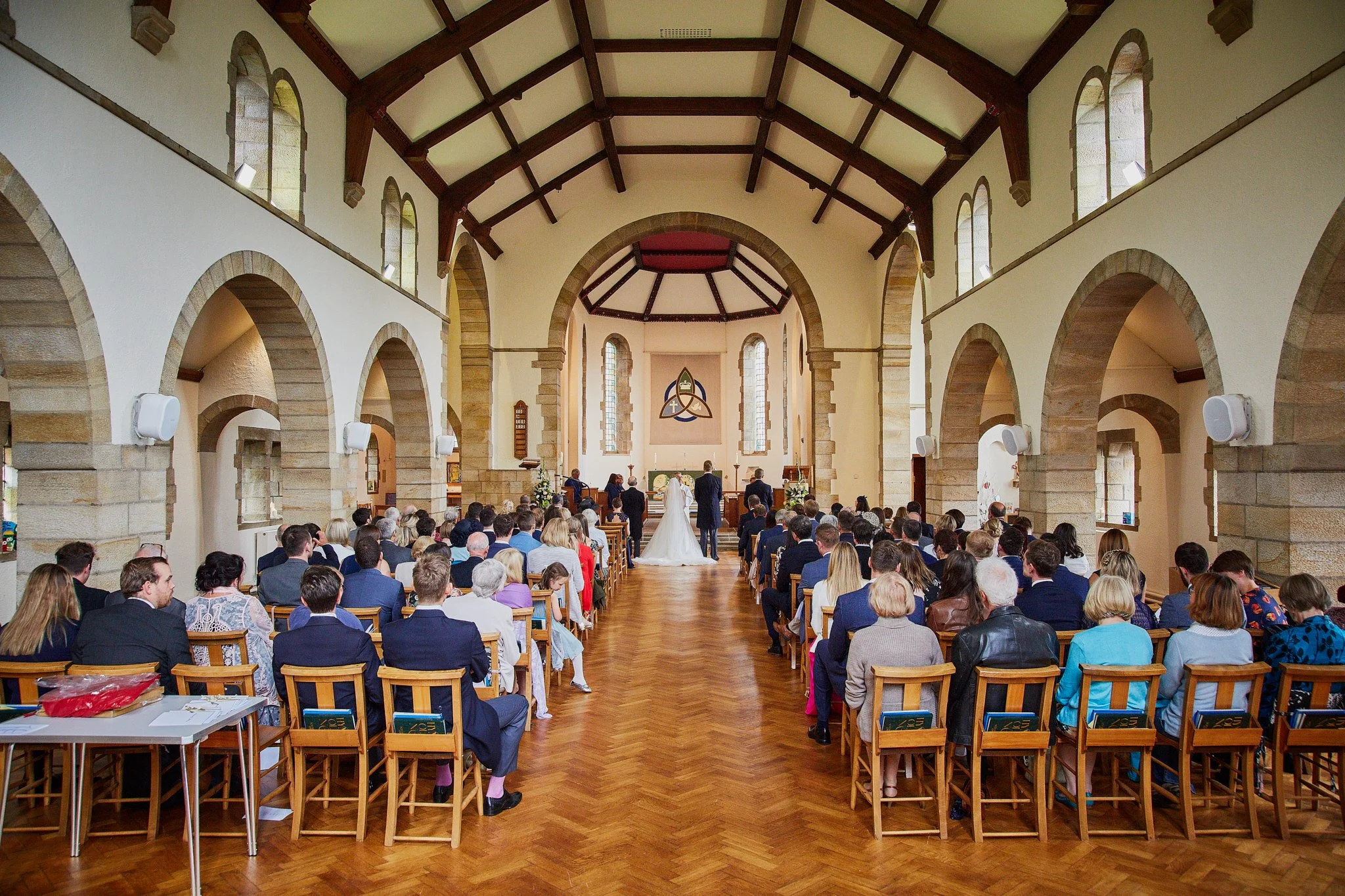 A wedding ceremony inside a church with a wooden floor, arched windows, and high vaulted ceiling. The bride and groom stand at the altar with a officiant, facing a seated congregation, during daytime.