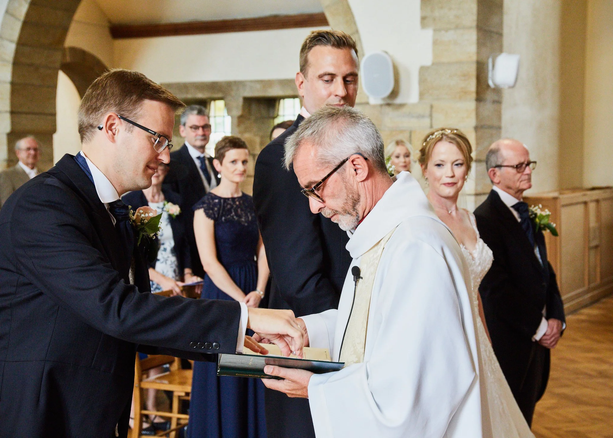 A wedding ceremony with a groom in a black tuxedo and a bride in a white dress, holding hands and exchanging rings inside a church, surrounded by guests.