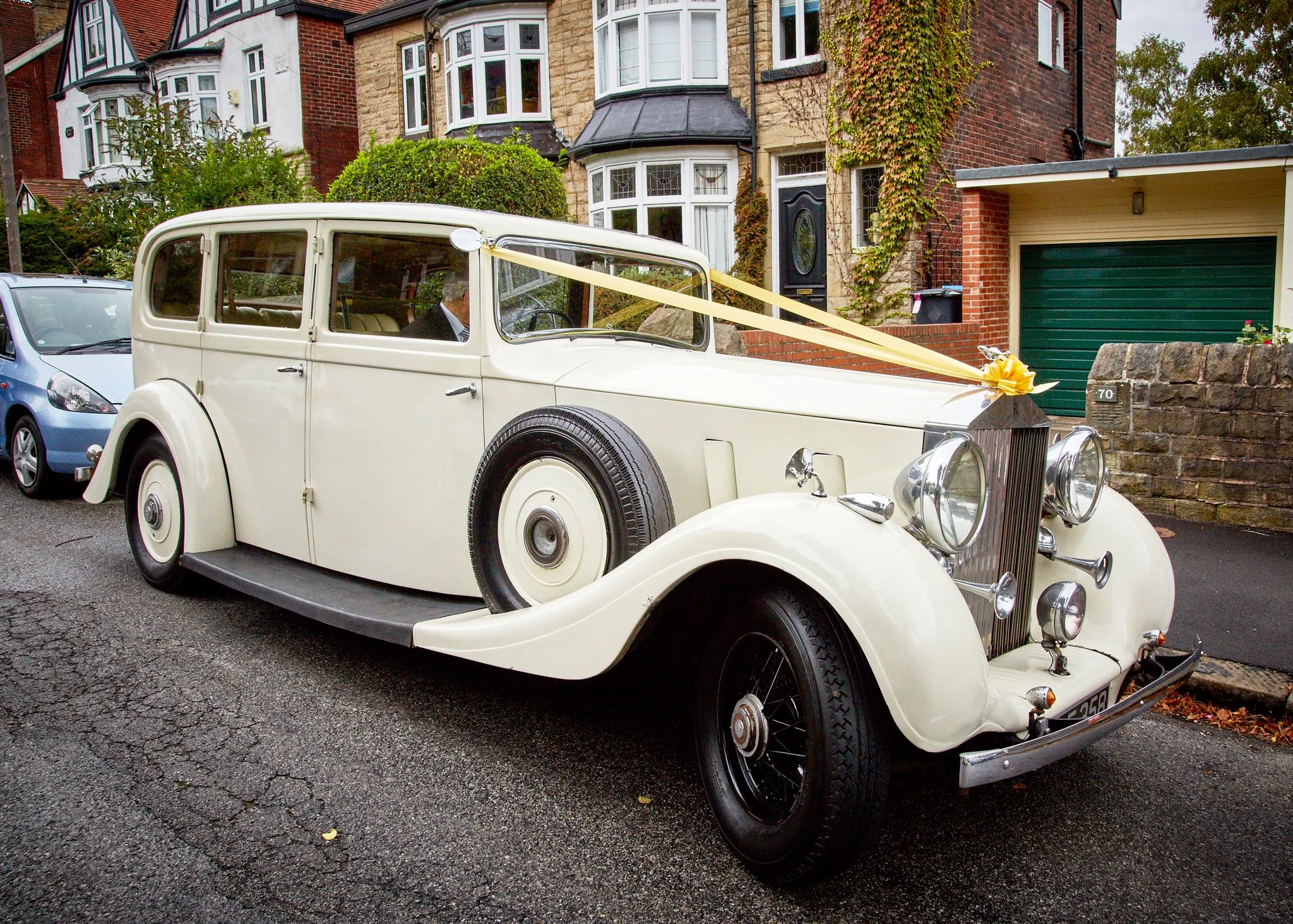 A vintage white car decorated with yellow ribbons parked in front of a brown brick house with a garage door. The house has ivy growing on the wall near the door and windows.