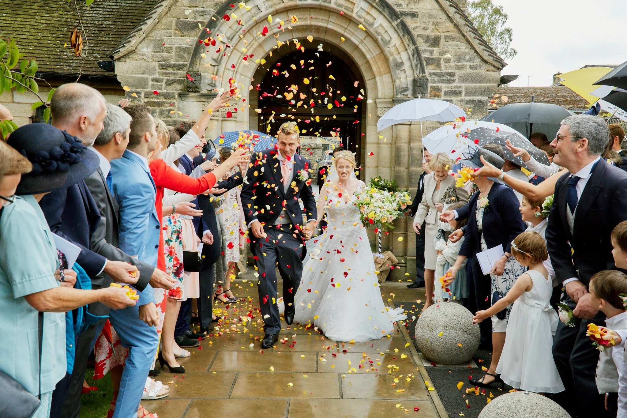 A bride and groom leaving a church while guests throw colorful flower petals. The scene is outdoors on a rainy day, with people holding umbrellas.