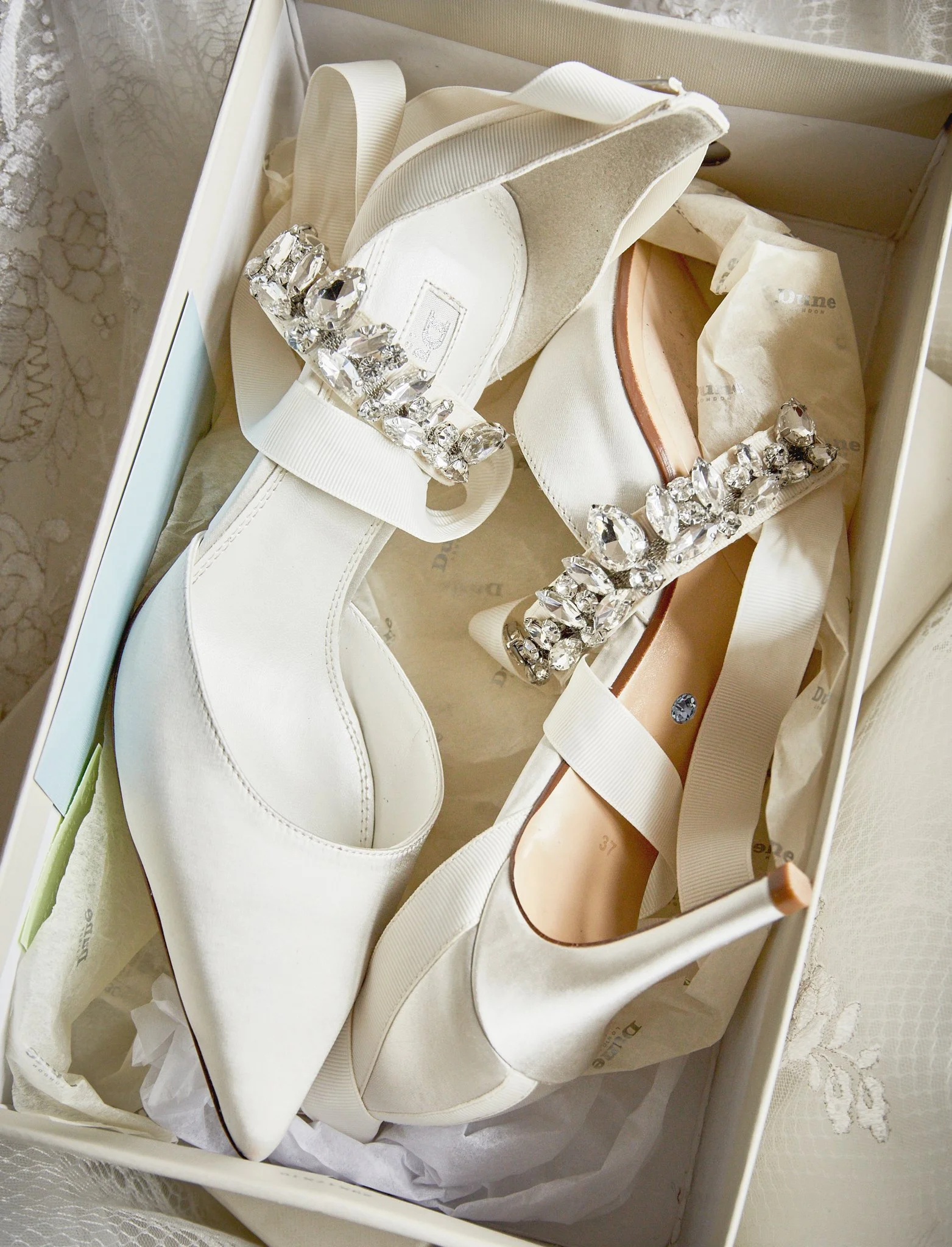 A pair of white high-heeled shoes with rhinestone embellishments on the straps, placed inside a white shoe box with tissue paper.