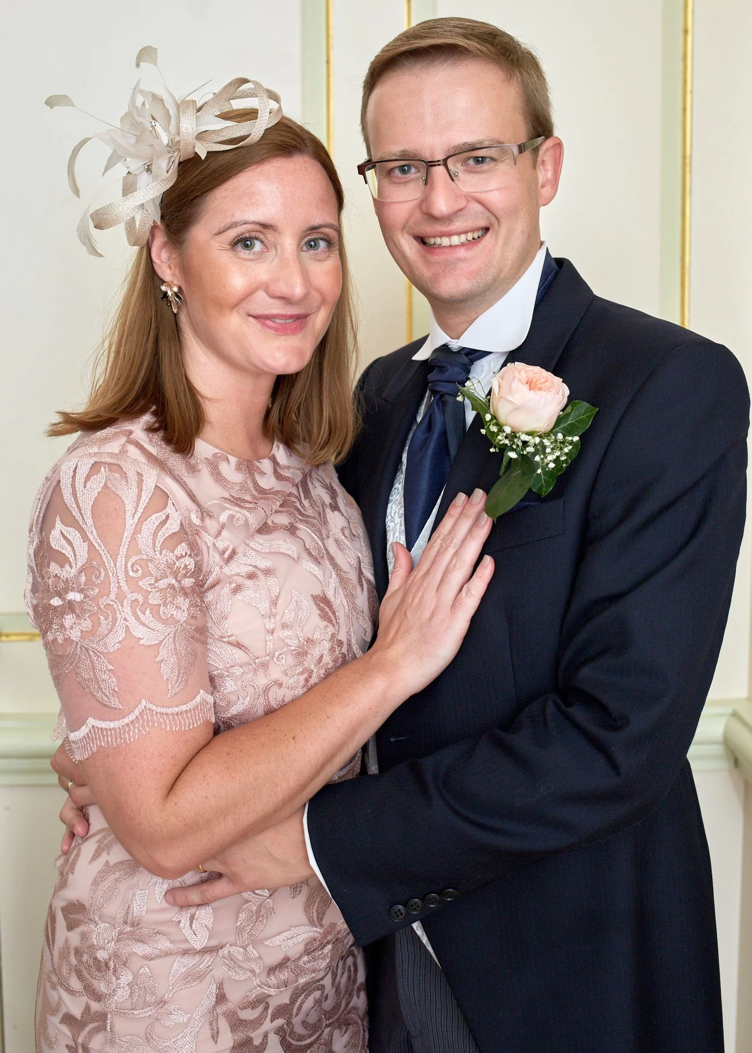 A woman and a man are smiling and posing for a photo in a formal setting. The woman has red hair, is wearing a pink dress with lace details, a fascinator hat, and earrings. The man has light brown hair, glasses, and is wearing a dark suit with a whit
