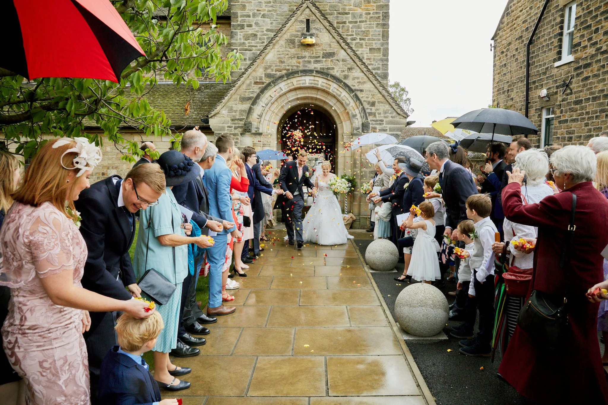 A newlywed couple walking through a crowd of guests during a wedding celebration outside a stone church, with colorful flower petals in the air and umbrellas held by guests on a rainy day.