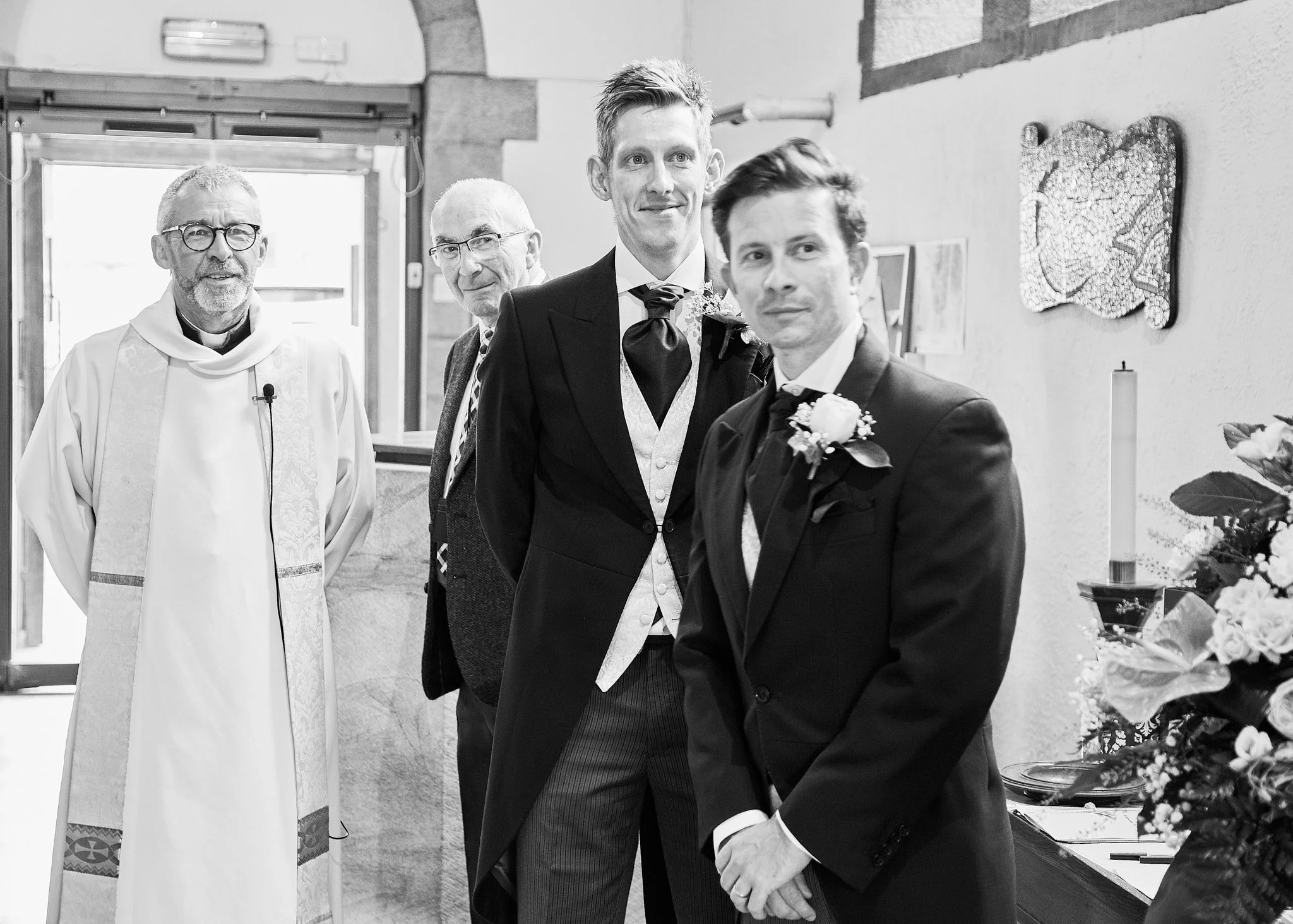 Four men dressed in suits and clerical attire standing together indoors, with some floral arrangements and artwork visible in the background.