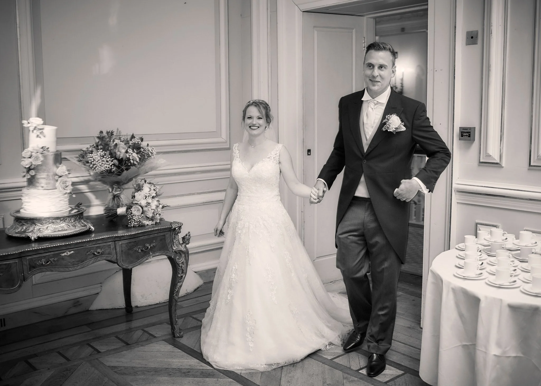A bride and groom holding hands at their wedding reception, with wedding cakes and floral arrangements nearby.