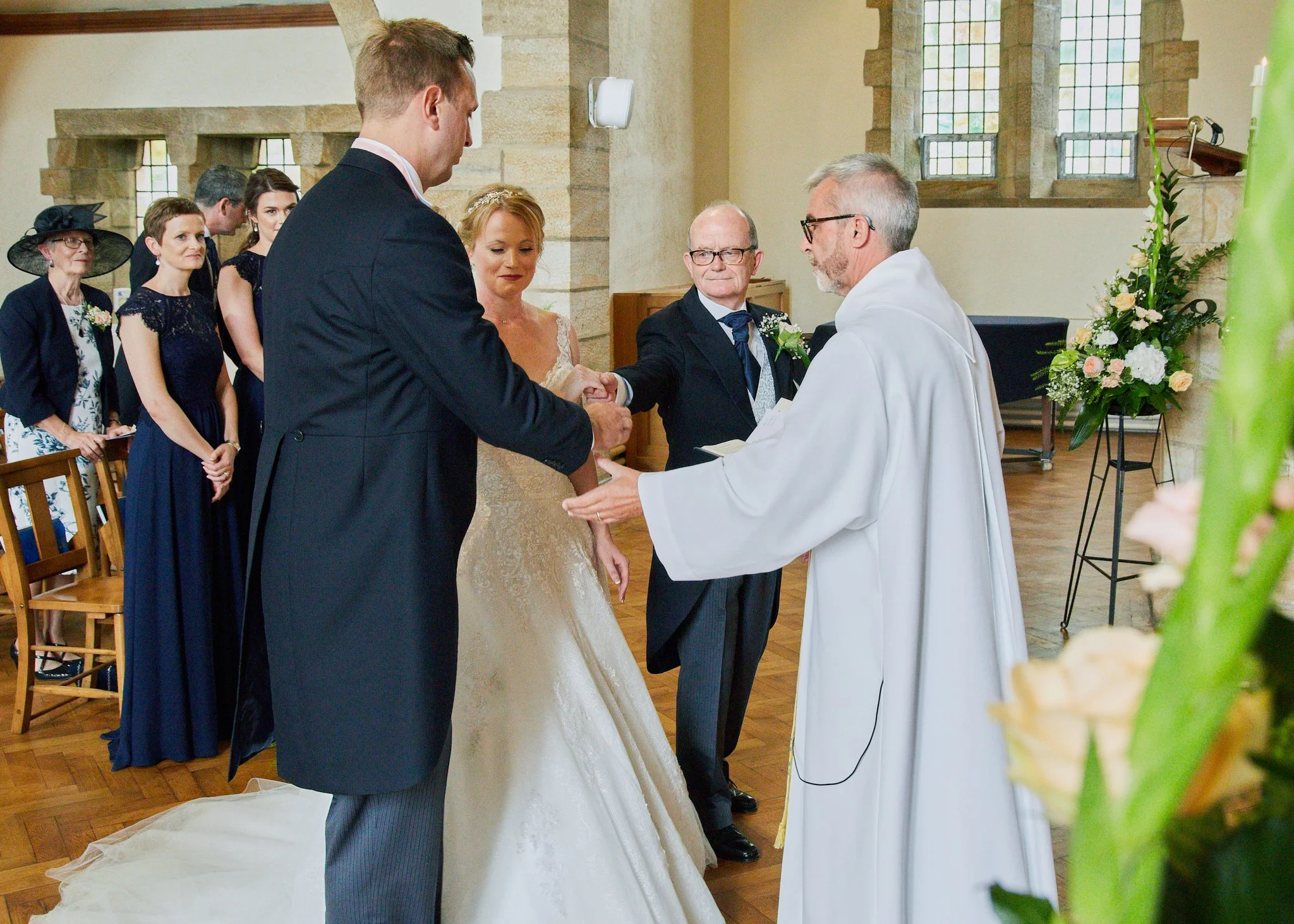 A bride and groom are holding hands during their wedding ceremony inside a church, with the priest officiating and guests in the background.