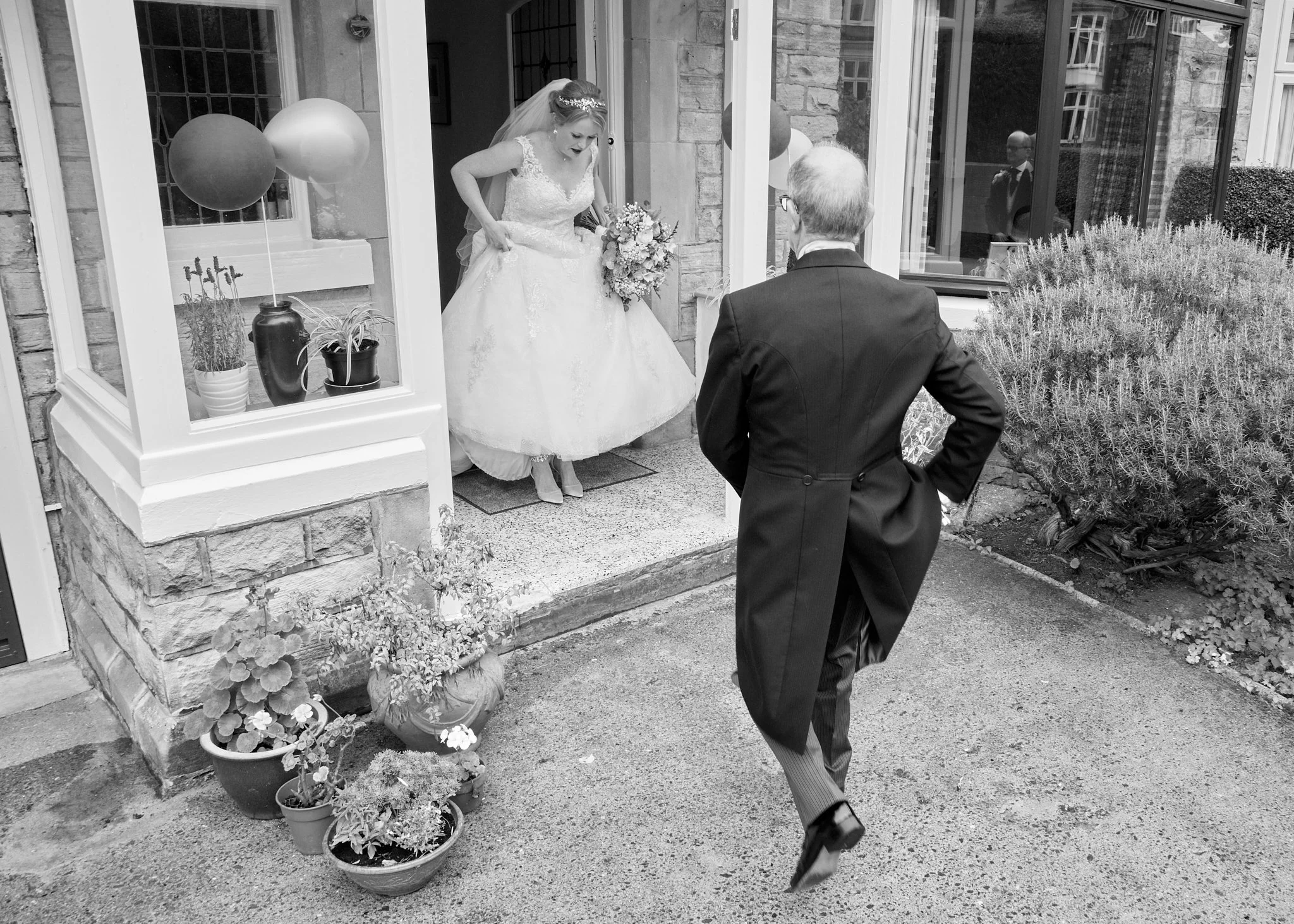 A bride in a wedding dress and veil holding a bouquet of flowers is standing at the doorstep of a house, looking down at a man in a suit who is stepping into the doorway with one foot raised. The scene is in black and white, with potted plants and ba