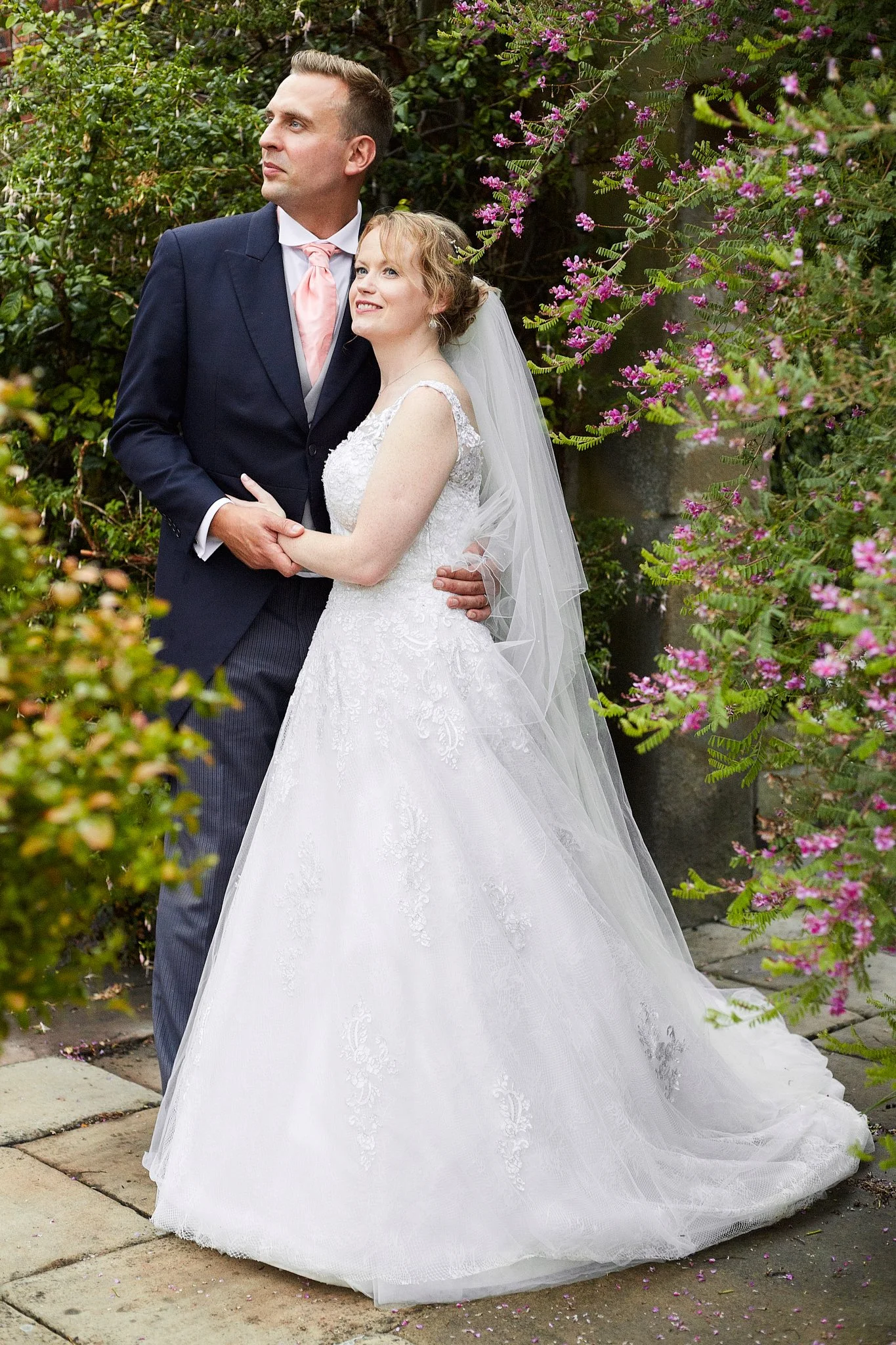 A bride and groom standing outdoors surrounded by flowering bushes. The bride is in a white wedding dress with a veil, and the groom is in a dark suit with a pink tie. They are holding hands and smiling softly, with the bride looking up and the groom