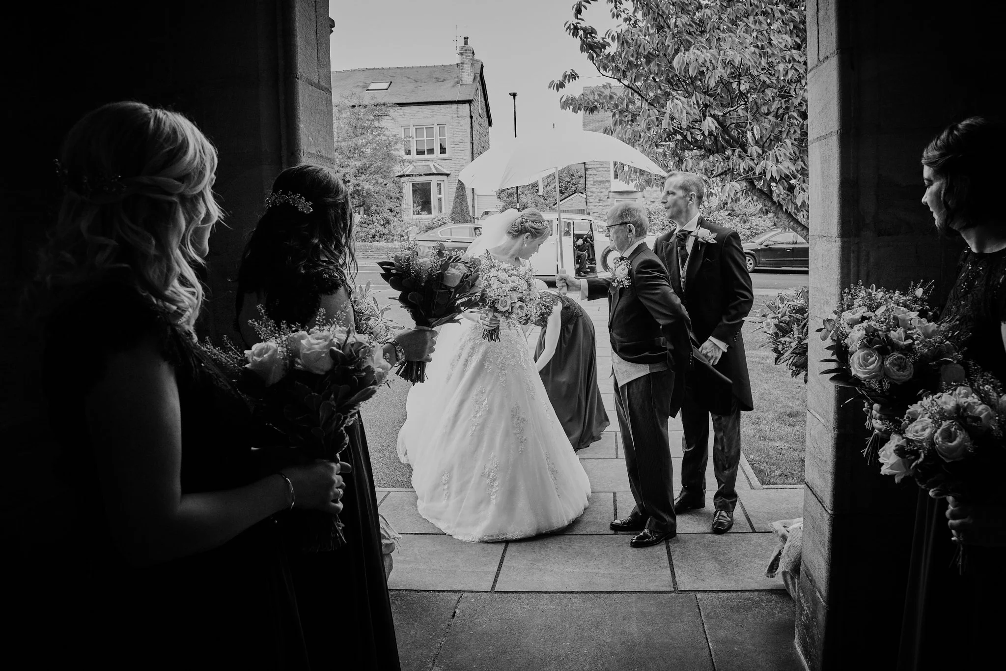 Black and white photo of a bride and groom exchanging vows outside, surrounded by bridesmaids and a woman holding flowers, under a large umbrella, with trees and houses in the background.