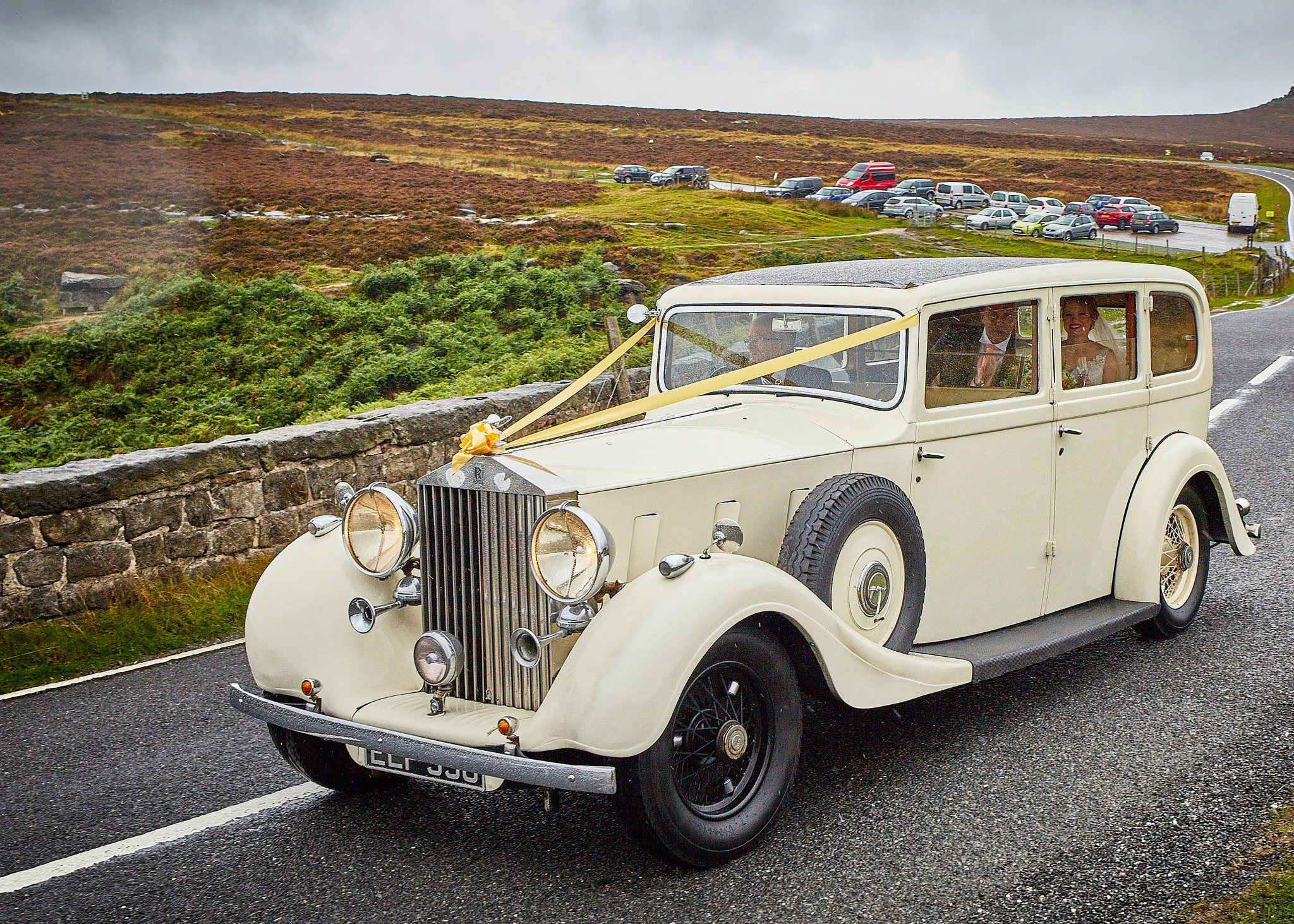 Vintage white car decorated for a wedding, parked on a wet road with a green and brown hilly landscape behind. Two women are inside the car, smiling, with wedding attire.