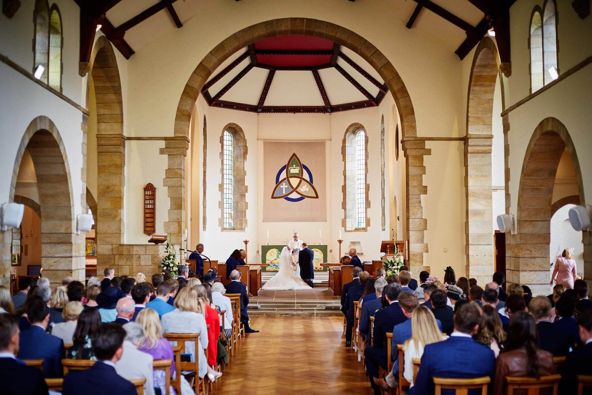 A wedding ceremony inside a church with a bride and groom standing at the altar with a priest officiating. The church interior features high stained glass windows, stone arches, and a congregation seated in wooden pews. The altar has religious symbol
