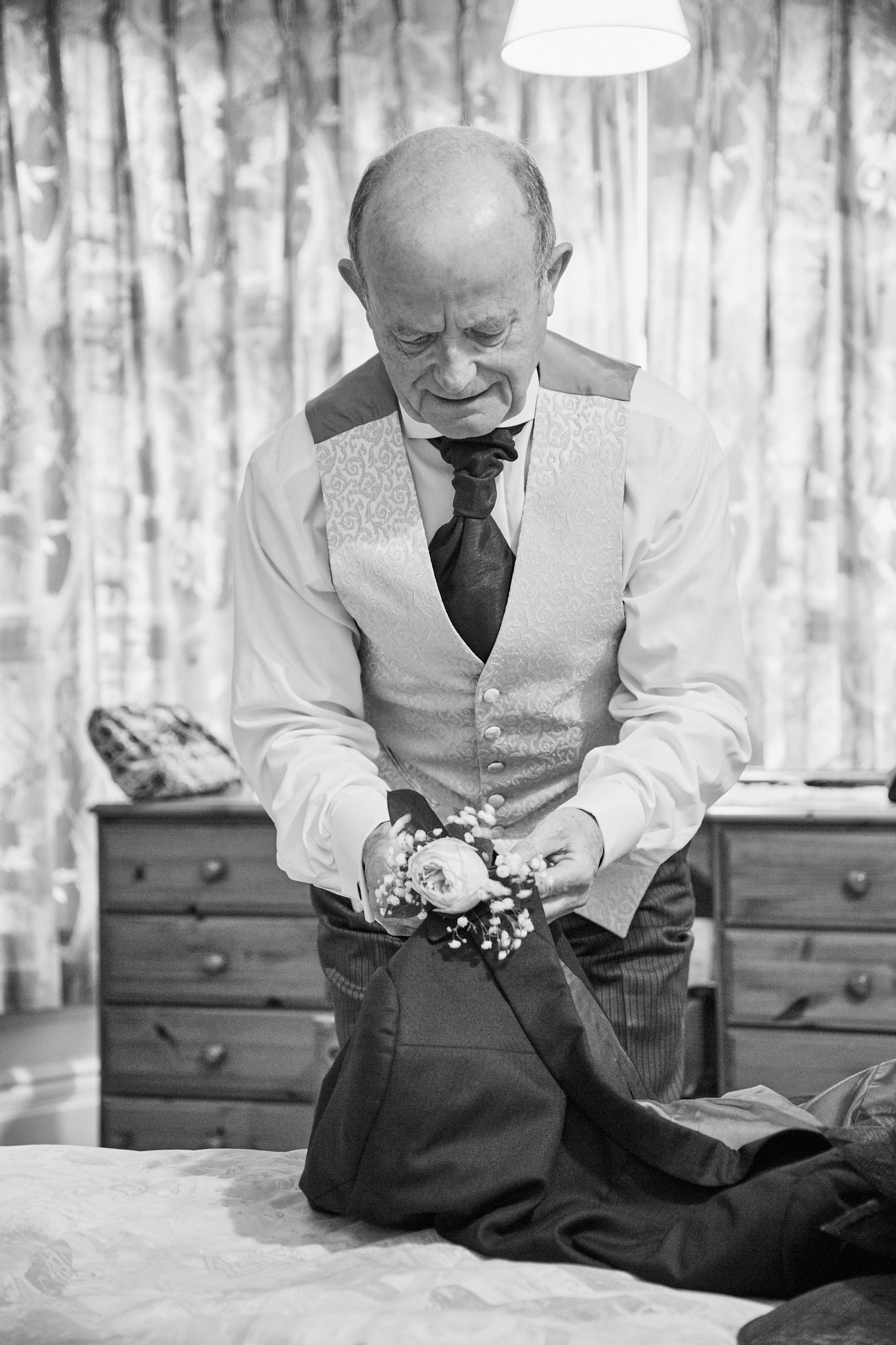 An elderly man with a bald head and a vest is holding a bouquet of flowers while kneeling in front of someone, in a room with curtains and a dresser in the background.