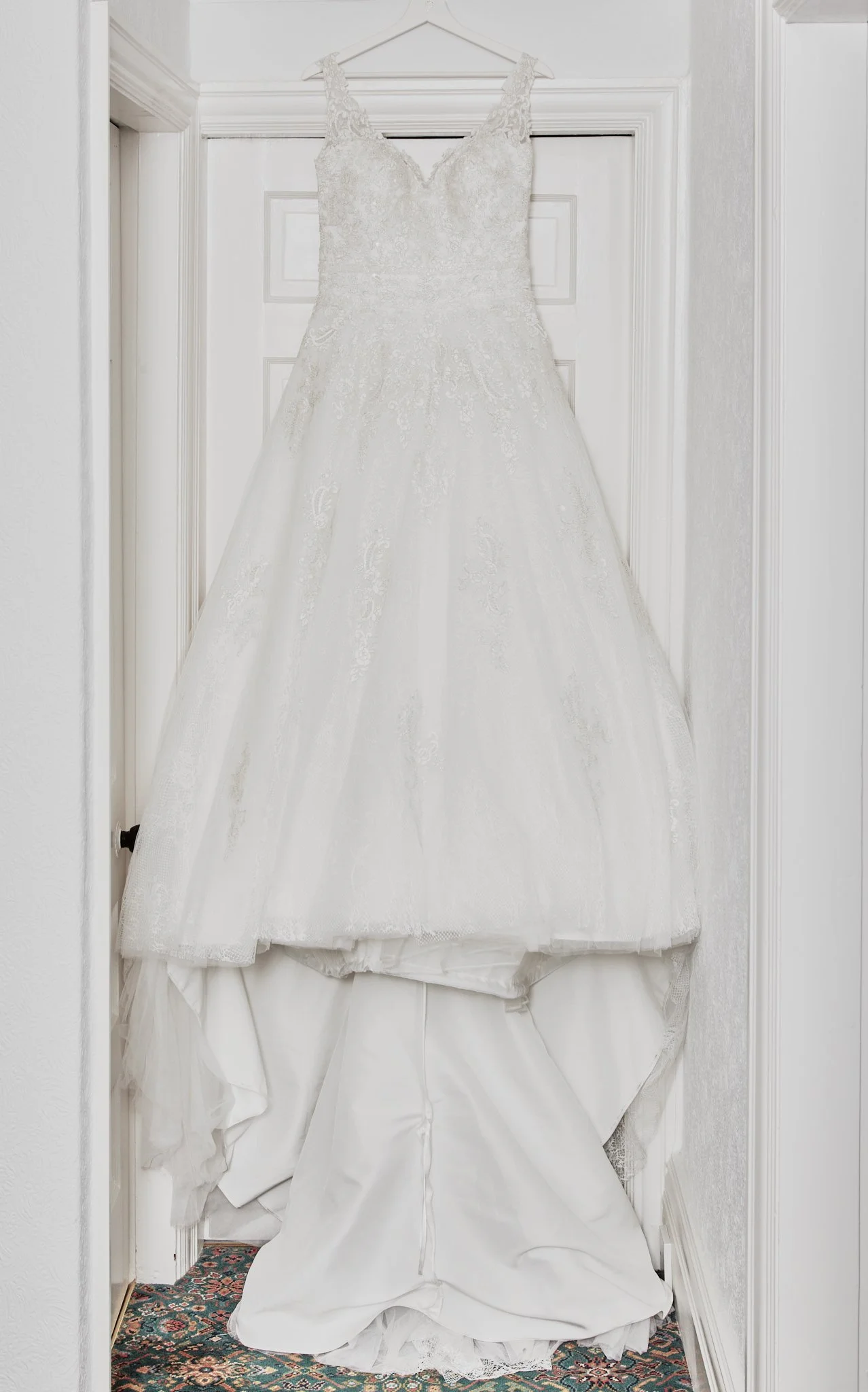 A wedding dress hanging on a white hanger on a door with ornate paneling, with a floral patterned rug visible at the bottom.