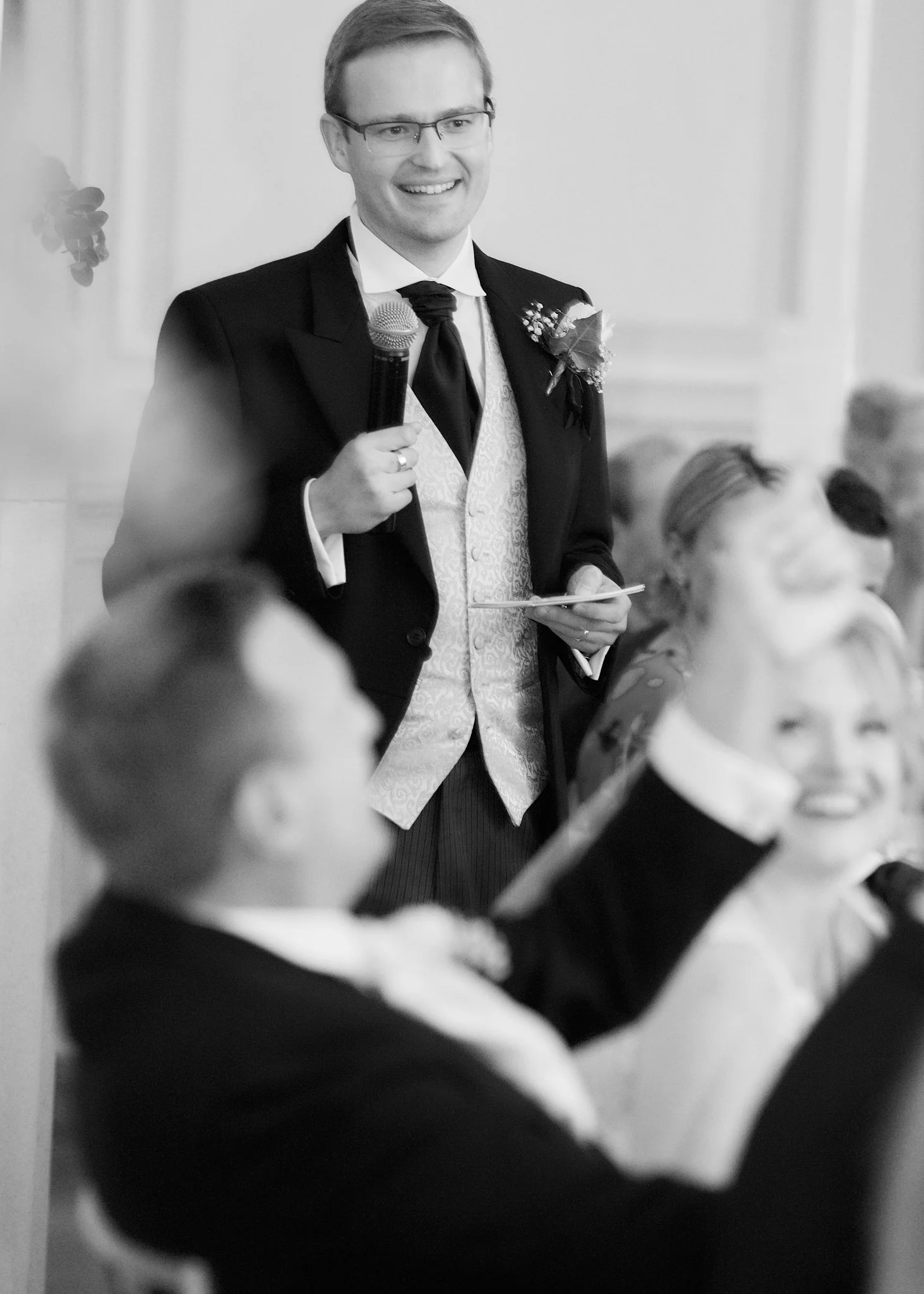 A man in formal attire, wearing glasses, holding a microphone, and smiling, at what appears to be a wedding reception or formal event. Other guests are blurred in the foreground.