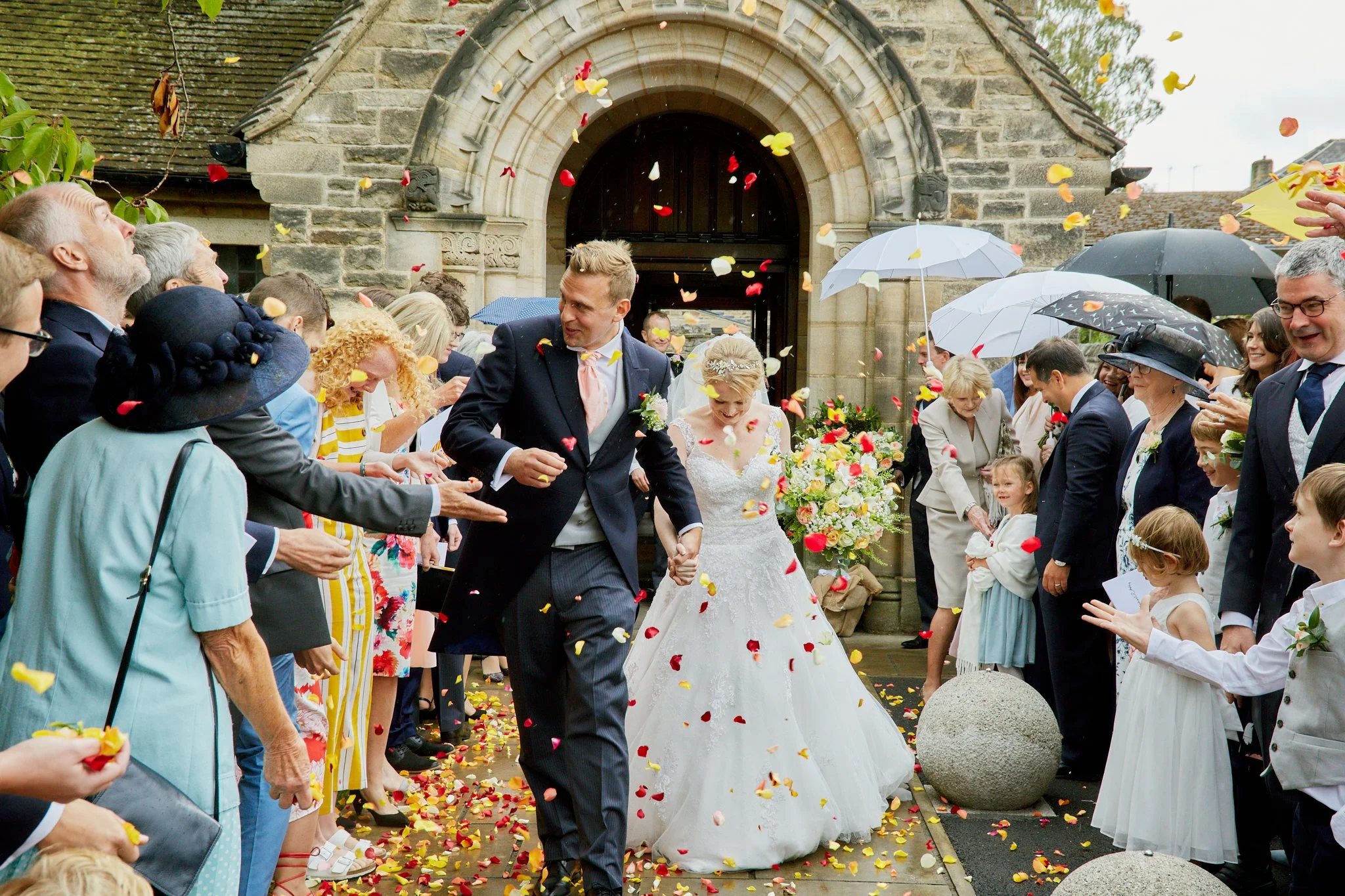 A newlywed couple walking through a crowd of friends and family, scattering flower petals outside a stone church.