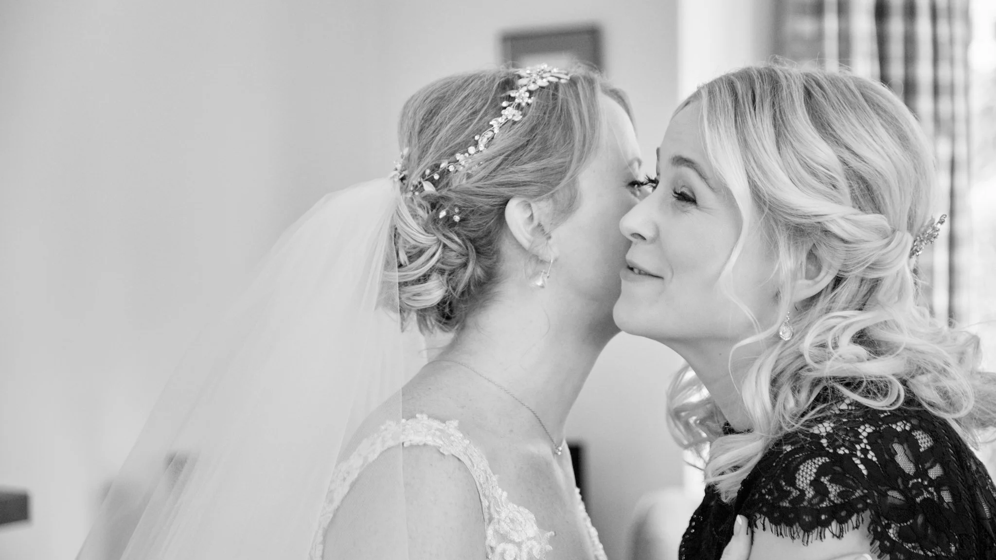 Black and white photo of a bride and another woman, likely her mother, touching noses and smiling inside a room.