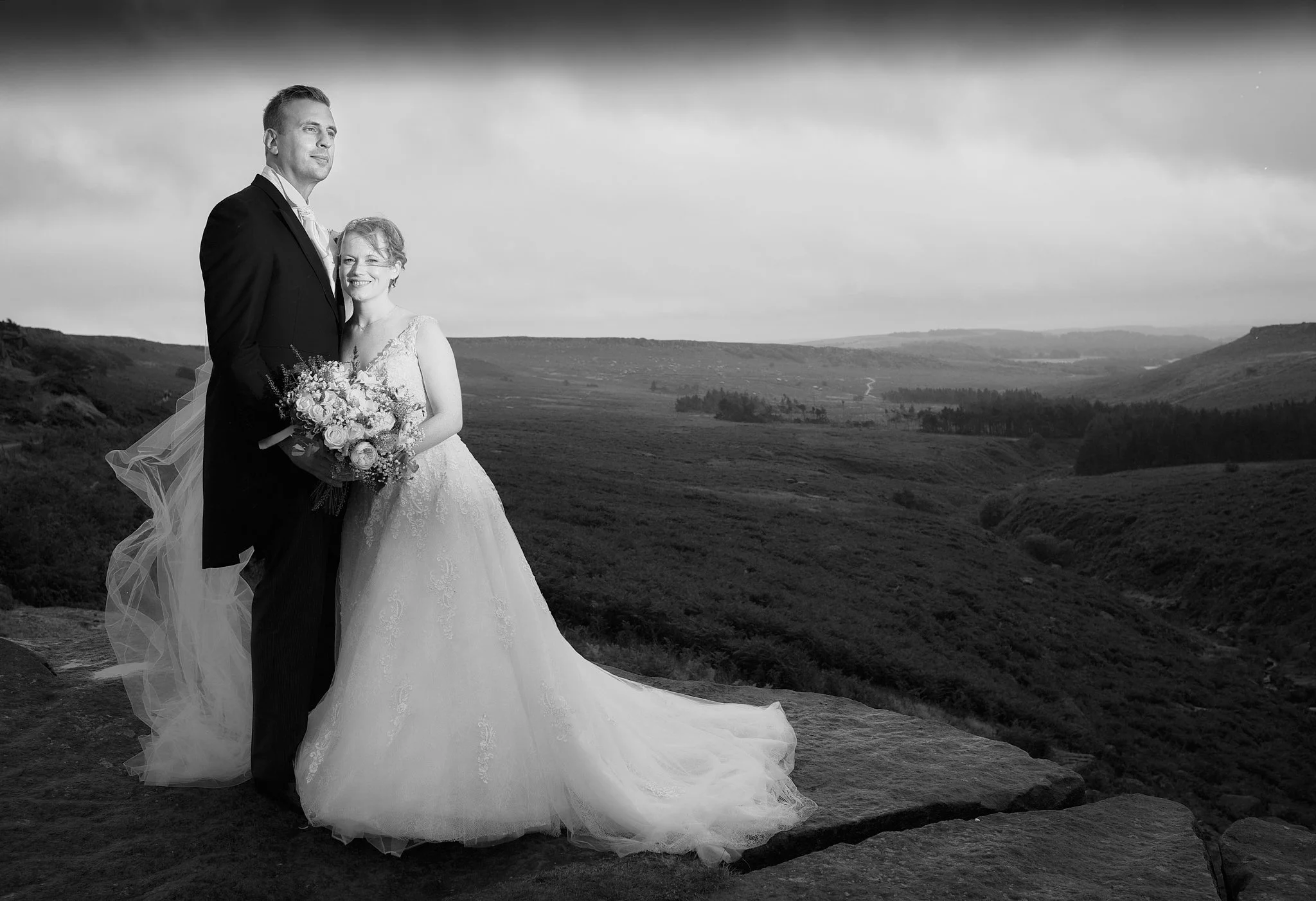Black and white photograph of a bride and groom standing outdoors on a rocky landscape with hills and a cloudy sky in the background. The bride is smiling and holding a bouquet, while the groom looks into the distance.