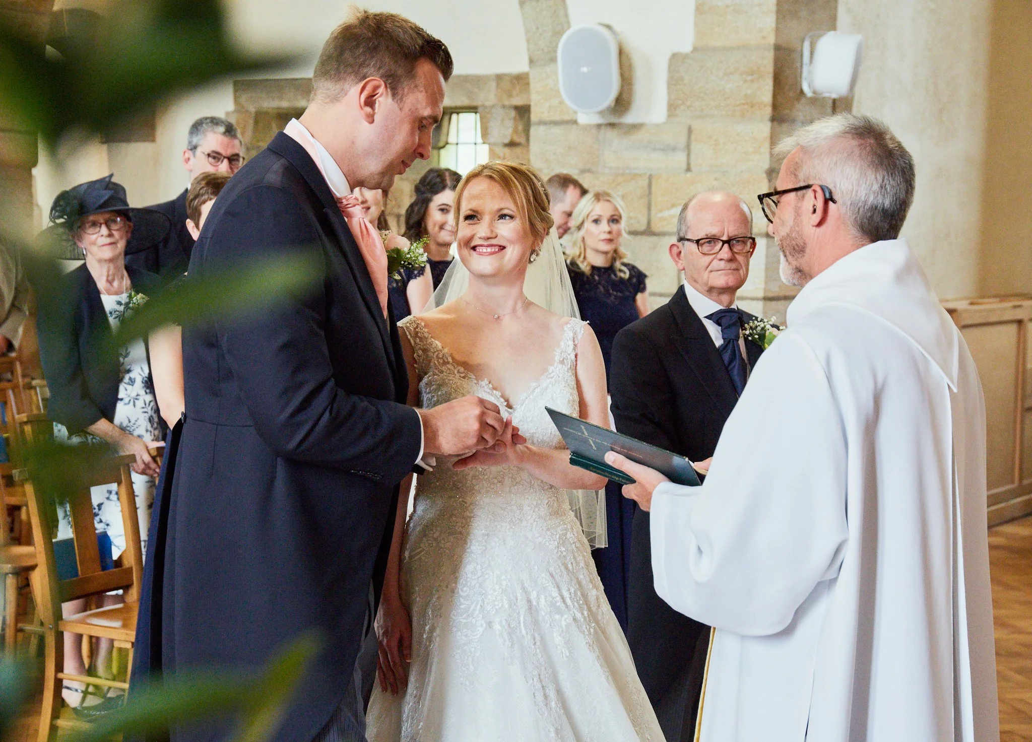 A couple getting married during a ceremony in a church, exchanging rings while an officiant oversees, with guests watching in the background.