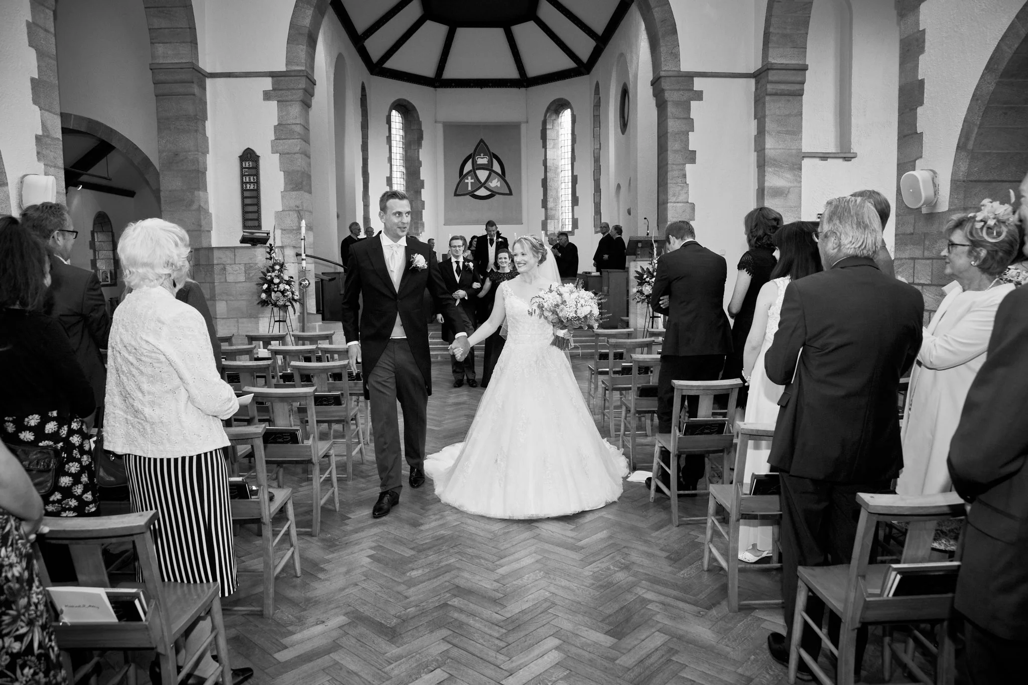 A bride and groom walking down the aisle in a church, surrounded by guests seated on wooden chairs, inside a church with stone walls, stained glass windows, and religious symbols.