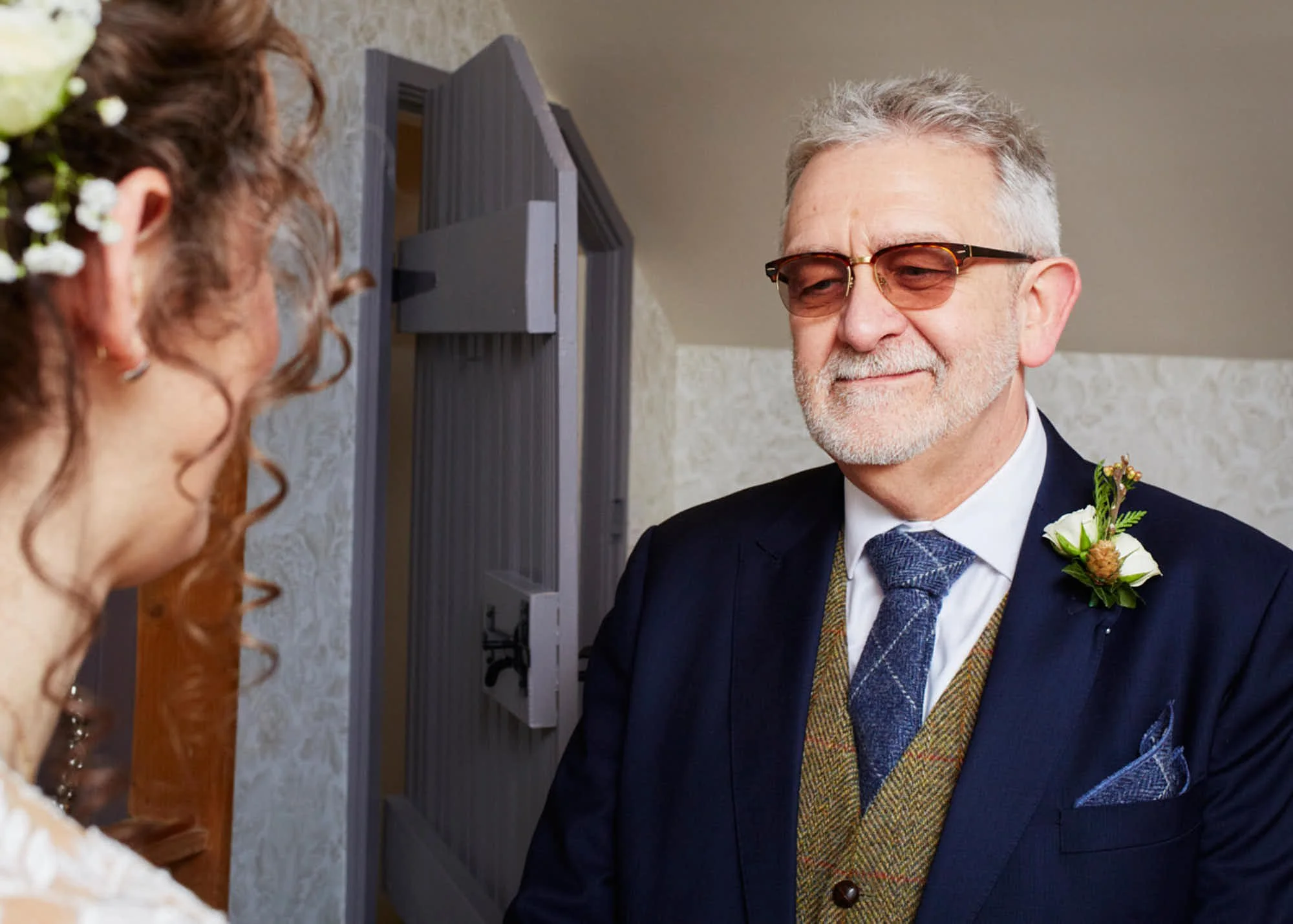 An older man with gray hair, glasses, and a beard in a dark suit and tie, smiling slightly, looking at a woman with curly red hair and a floral hair accessory.