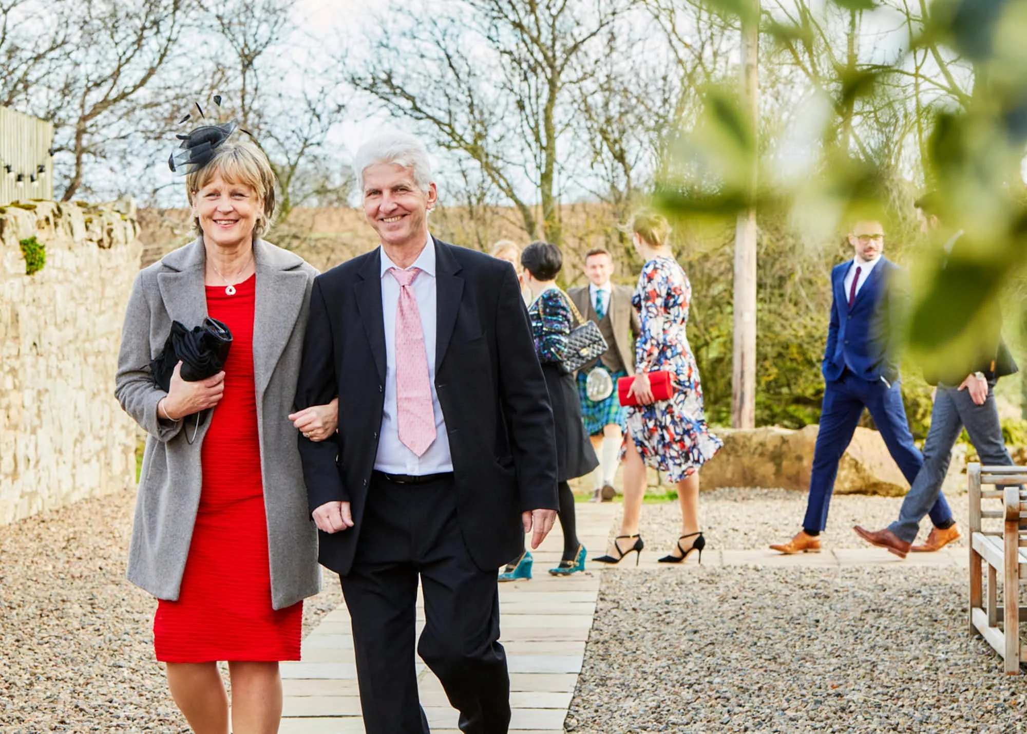 Smiling woman in red dress with gray coat and fascinator hat walking arm in arm with smiling man in black suit and pink tie at an outdoor event, with other people dressed formally in the background.