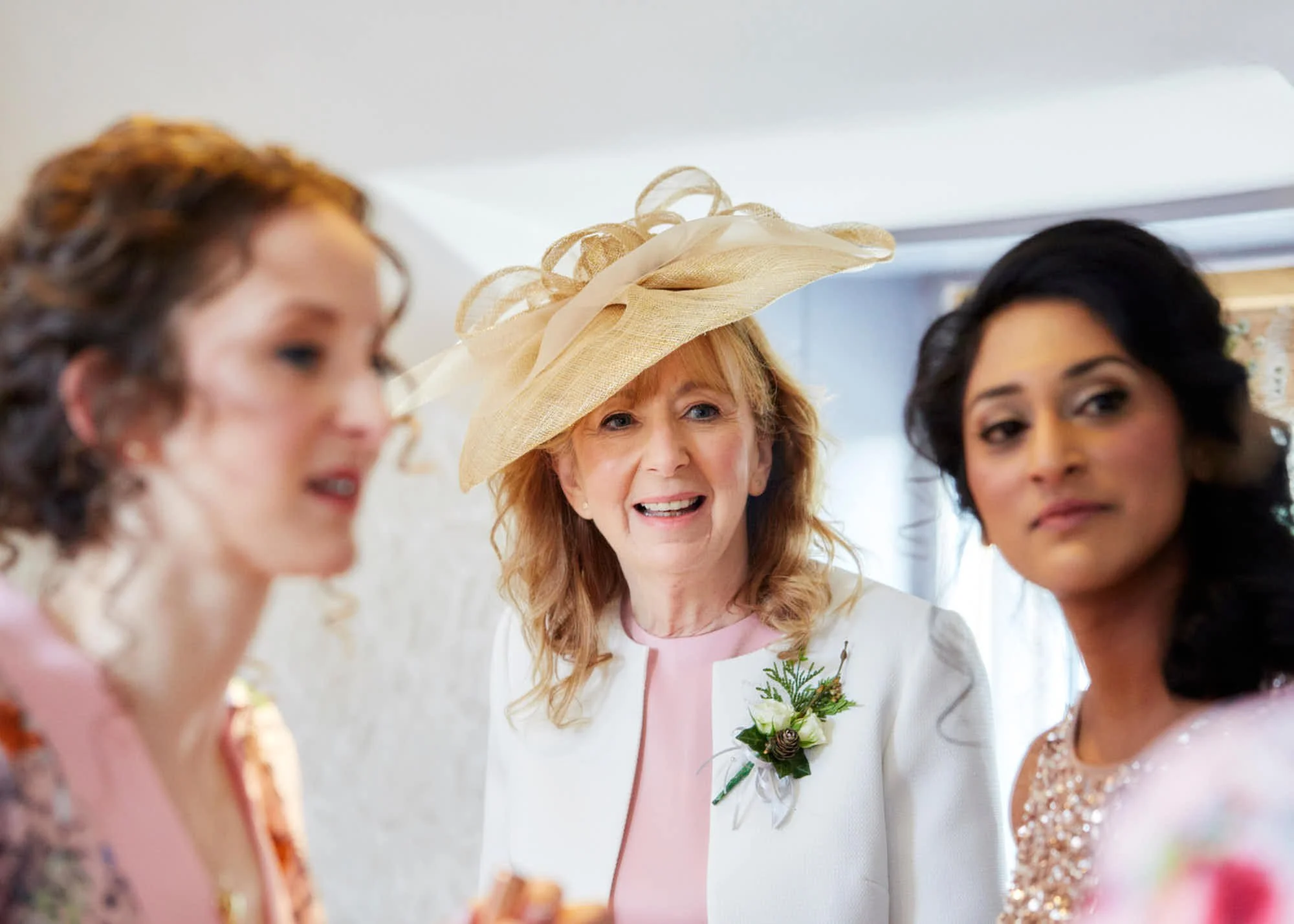 A woman wearing a large, beige, decorative hat is among a group of women at a social event, with some wearing formal attire and elegant dresses.