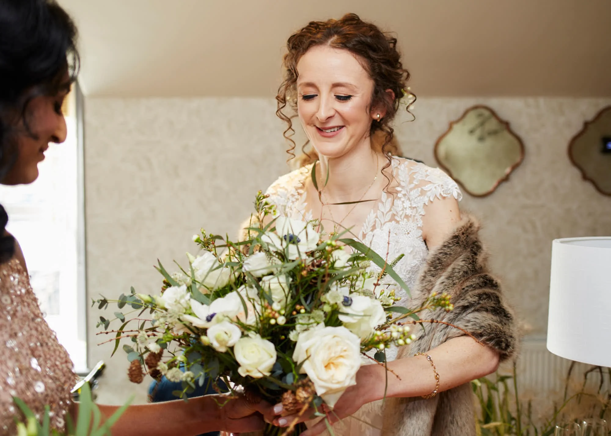 A woman with curly hair, wearing a white lace dress, holds a bouquet of white flowers and greenery, smiling as she looks at it.