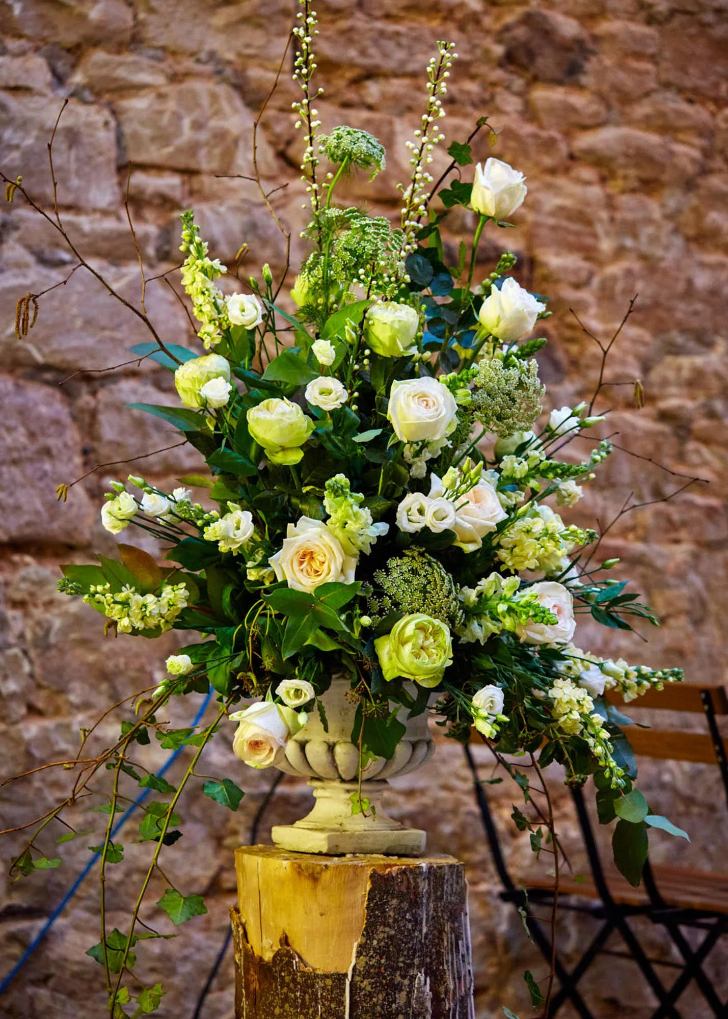 A floral arrangement with white roses, green foliage, and delicate white flowers in a cream-colored urn on a wooden stand against a brick wall background.