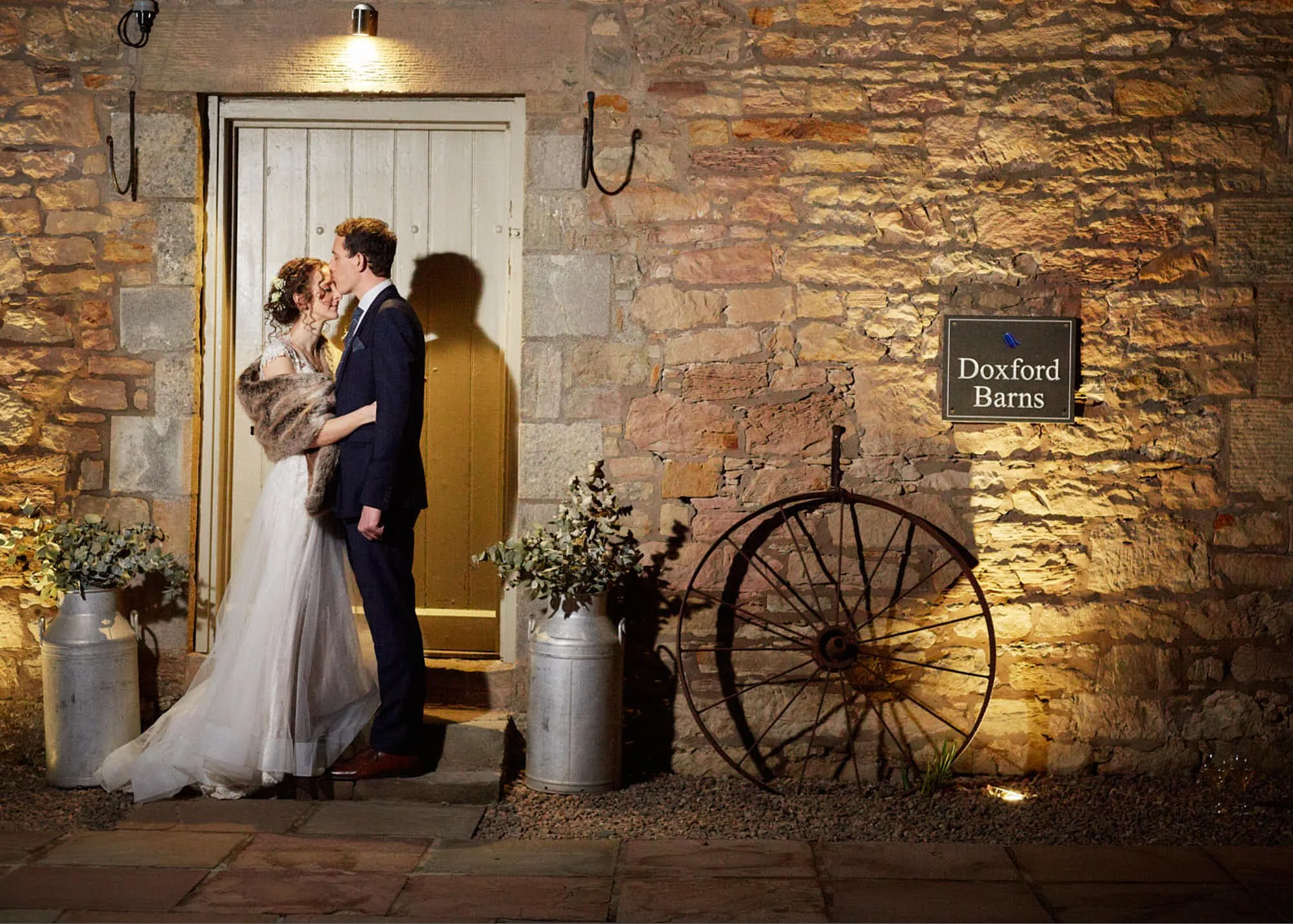 A bride and groom sharing a kiss in front of a rustic brick wall with a sign that reads "Doxford Barns", decorated with plants and vintage wheel decor, soft ambient lighting, and floral arrangements.