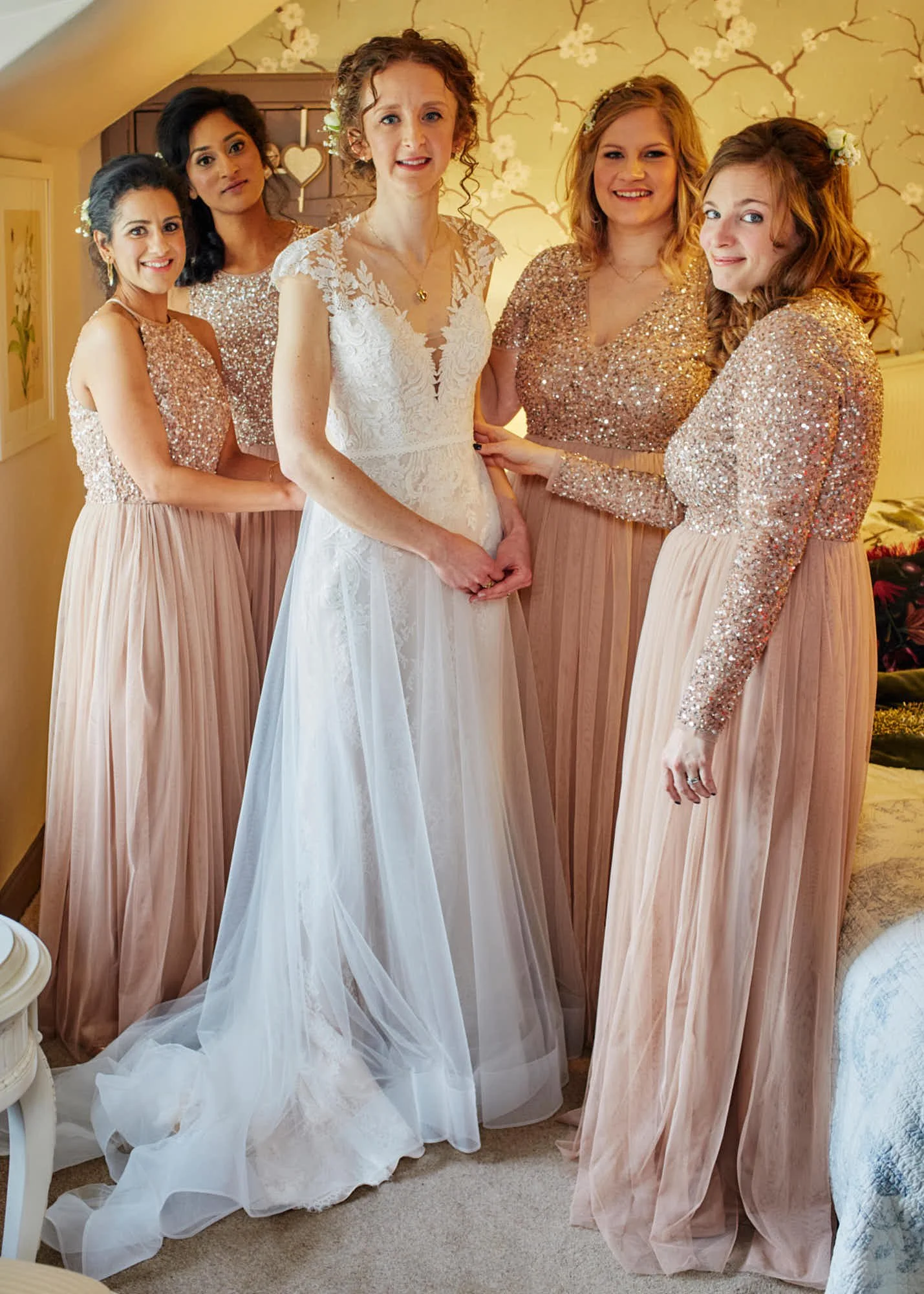A bride with curly red hair wearing a white lace wedding gown stands with five bridesmaids in blush pink, glittery long-sleeve tops and matching long skirts, inside a room with floral wallpaper, a white door, and a heart-shaped decoration in the back