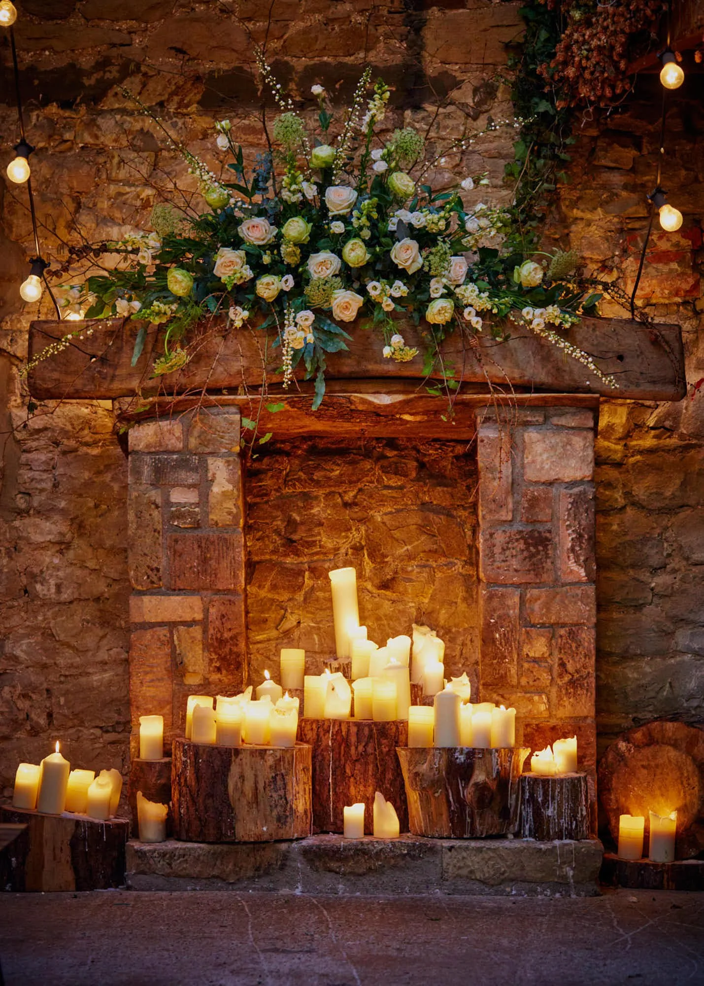 A stone fireplace decorated with a large, lush bouquet of white and pale pink roses, greenery, and branches at the top. Several lit candles are arranged on the hearth and in front of the fireplace, creating a warm, cozy atmosphere.