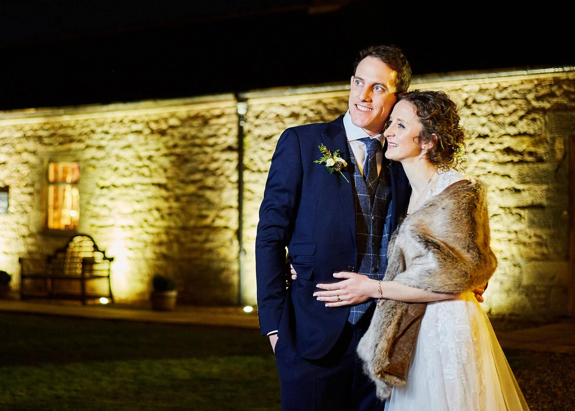 A wedding couple standing close together outdoors at night, with a brick wall illuminated by warm lighting behind them. The groom is wearing a dark suit with a boutonniere, and the bride is in a white lace dress with a fur shawl.