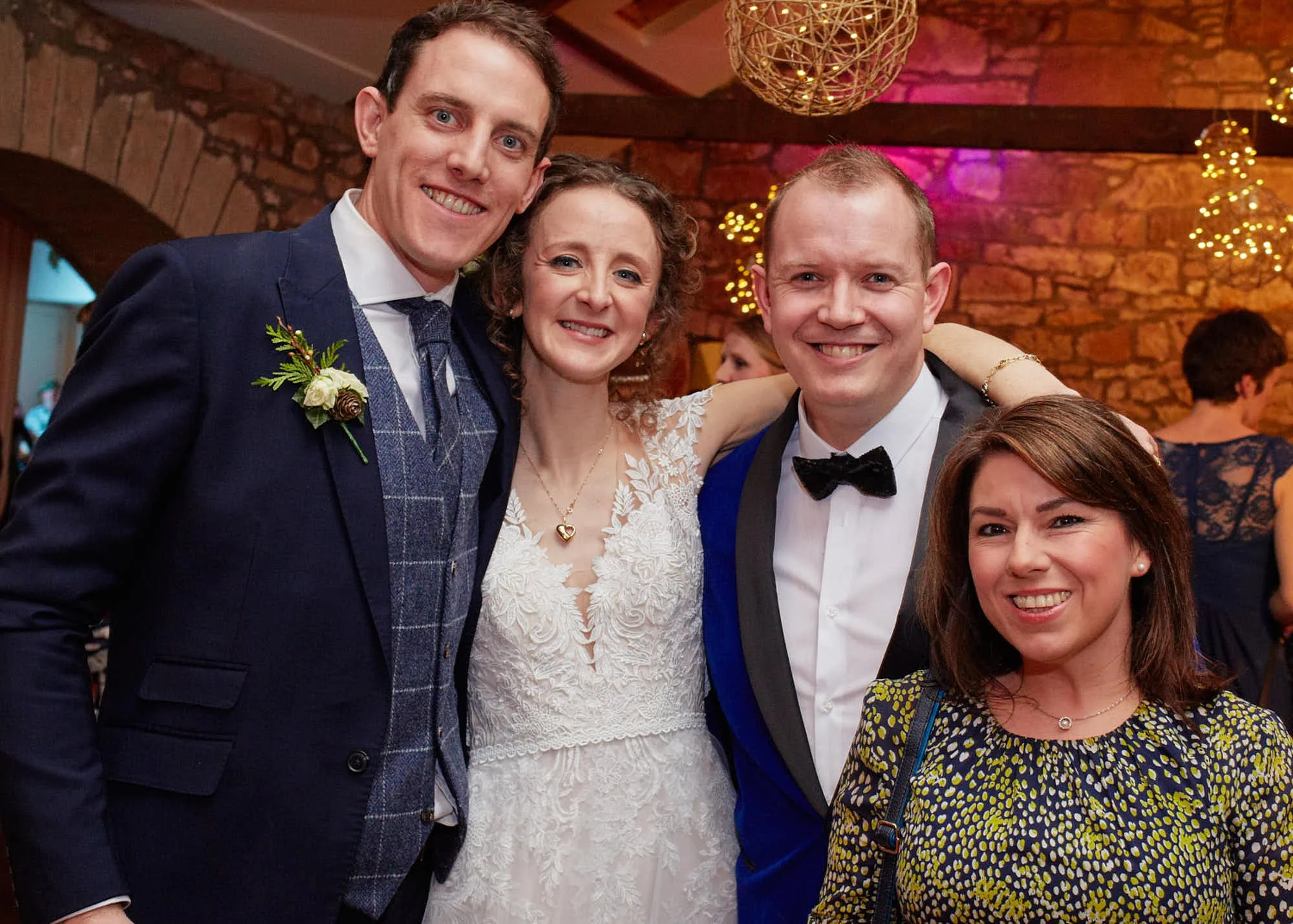 Four people at a wedding, smiling at the camera in a warmly lit venue with brick walls and string lights.